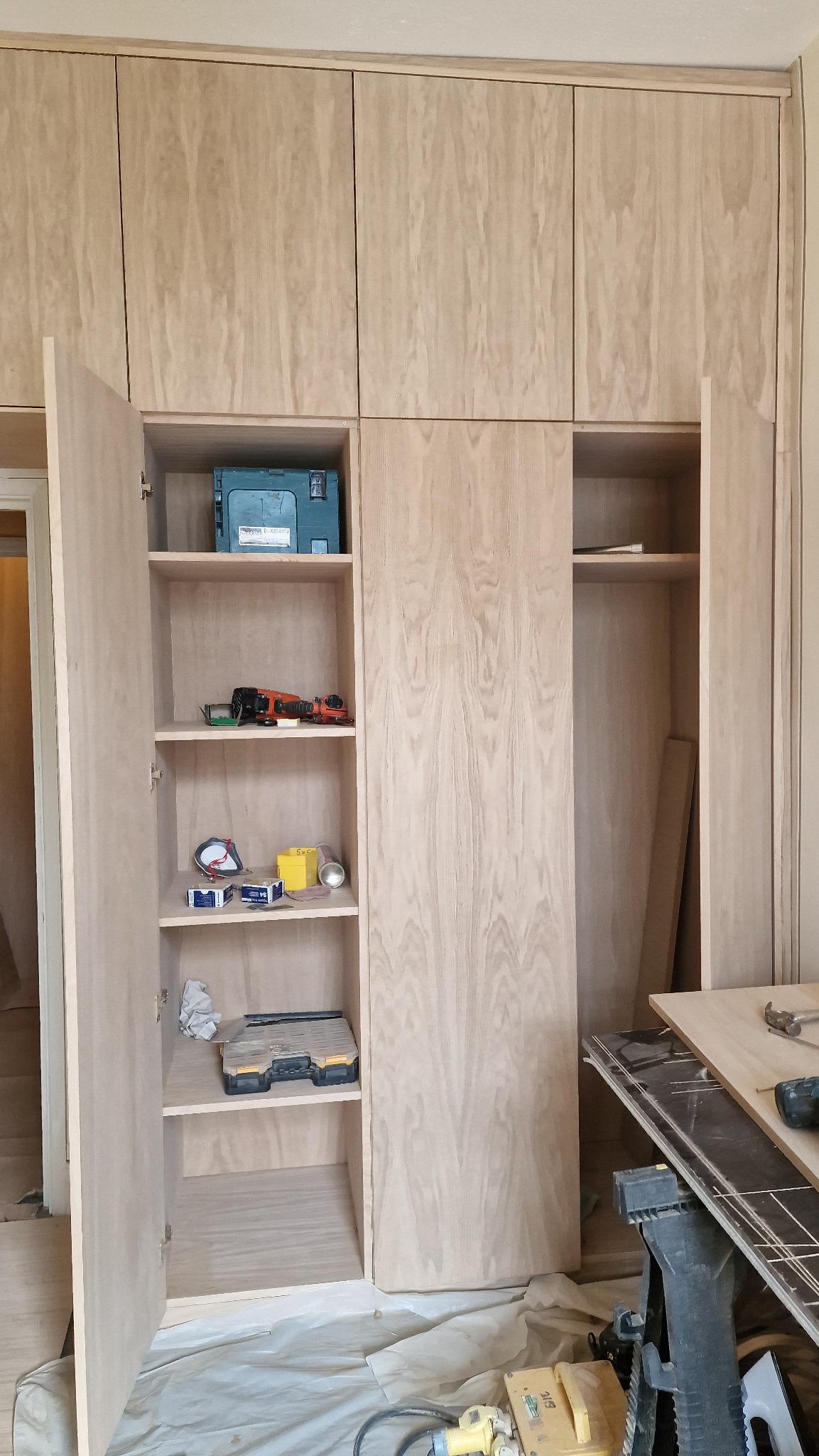 Wnętrze szafy wnękowej z jasnego drewna w trakcie montażu, widoczne narzędzia i akcesoria na półkach, zabezpieczona podłoga.