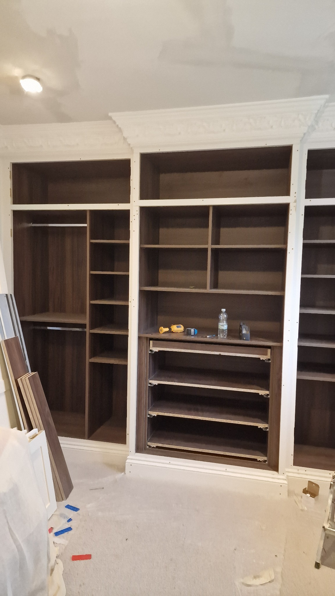 Wnętrze szafy wnękowej w trakcie montażu, widoczne półki, drążki, narzędzia i materiały wykończeniowe na podłodze, białe listwy dekoracyjne u góry.