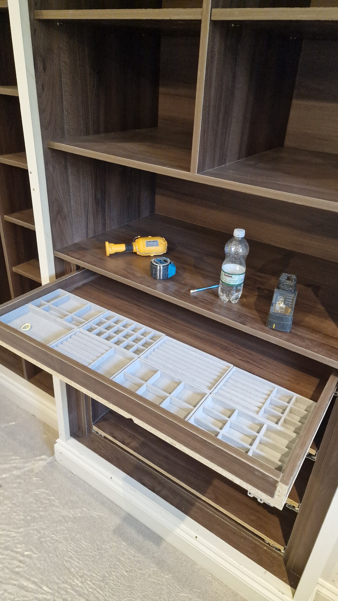 Wysuwana szuflada z przegródkami na biżuterię w brązowej szafie wnękowej, obok narzędzia, miarka i butelka wody na półce.