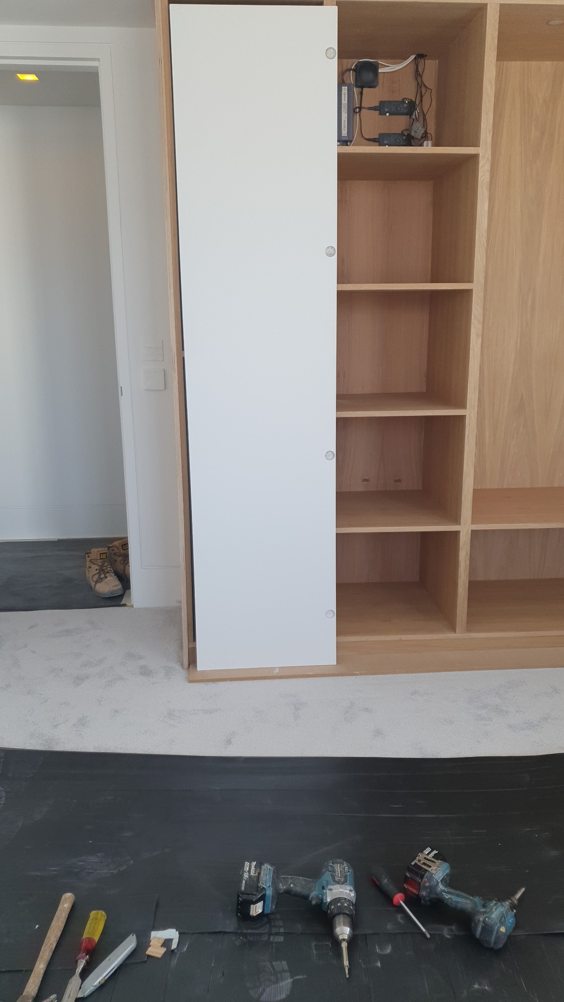 Montaż szafy wnękowej z przesuwnymi, białymi drzwiami; widoczne narzędzia na czarnej folii zabezpieczającej podłogę, częściowo otwarte półki z okablowaniem wewnątrz.