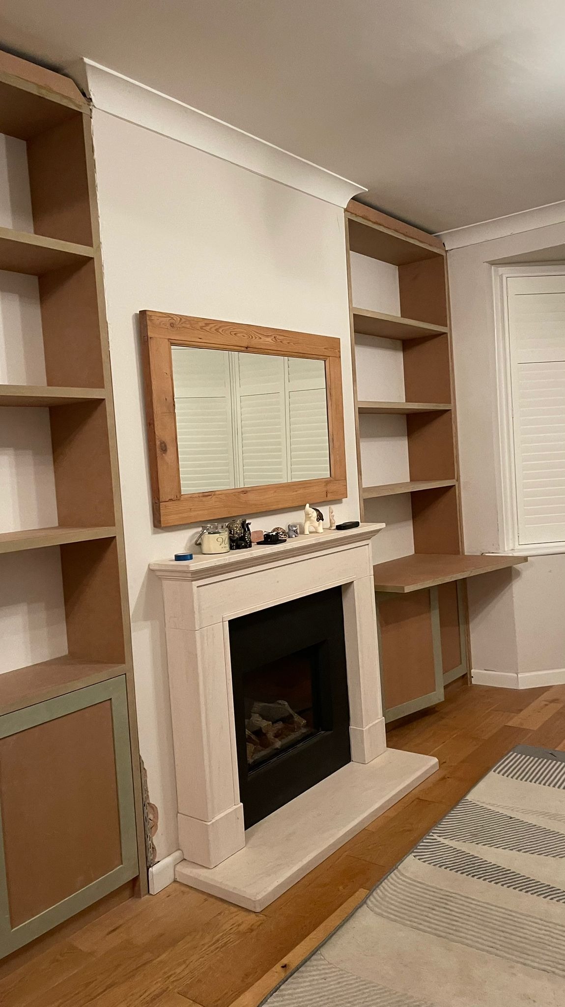 Wnętrze pokoju w trakcie montażu szafy wnękowej z półkami, wbudowanej po bokach kominka z drewnianą ramą lustra, widoczne niedokończone wykończenia i surowe płyty meblowe.