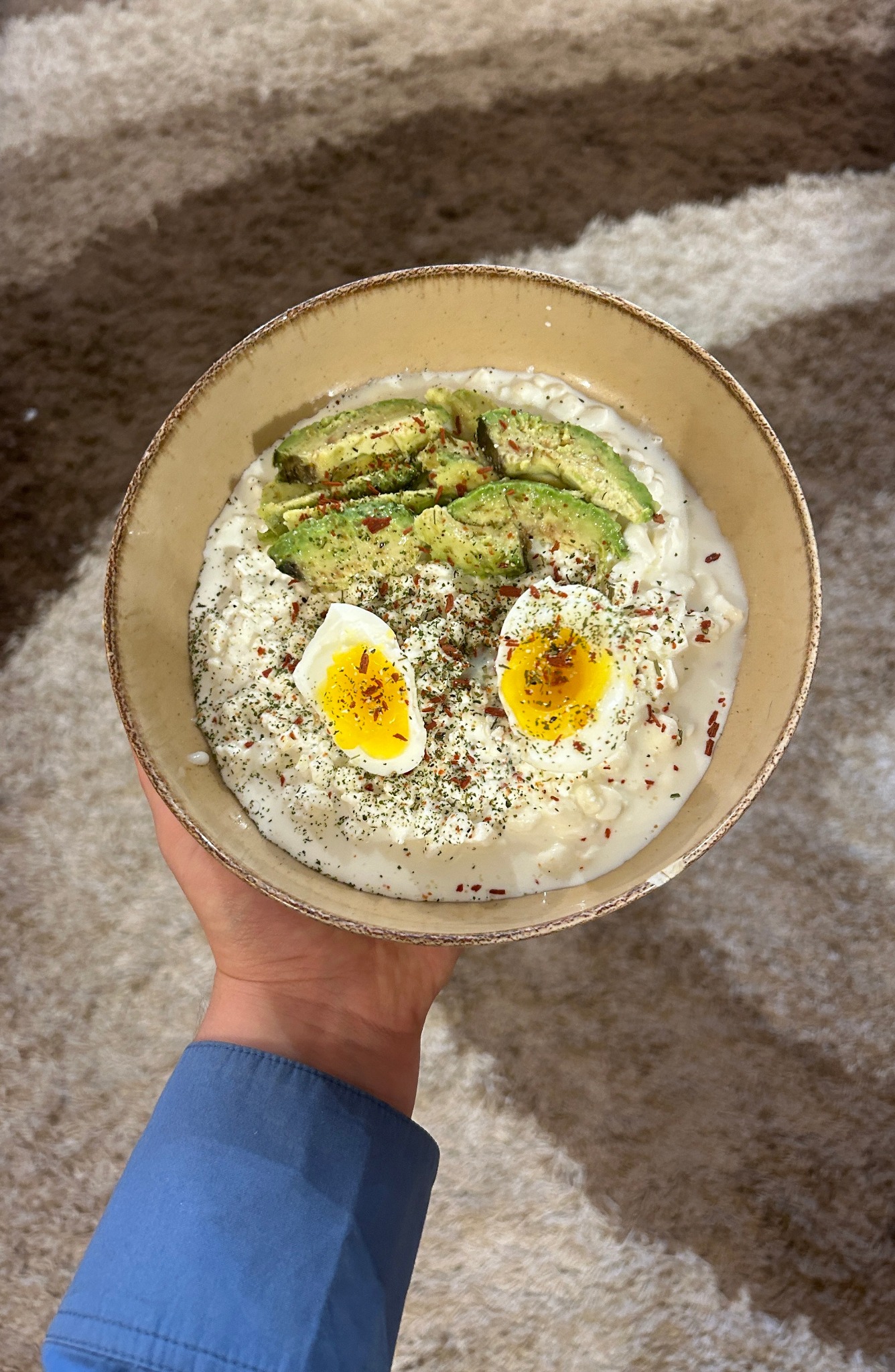 Kremowa owsianka z dodatkiem awokado, dwóch przekrojonych na pół jajek na miękko, posypana zielonymi ziołami i czerwonym pieprzem, trzymana w dłoni na tle beżowego dywanu z długim włosiem.