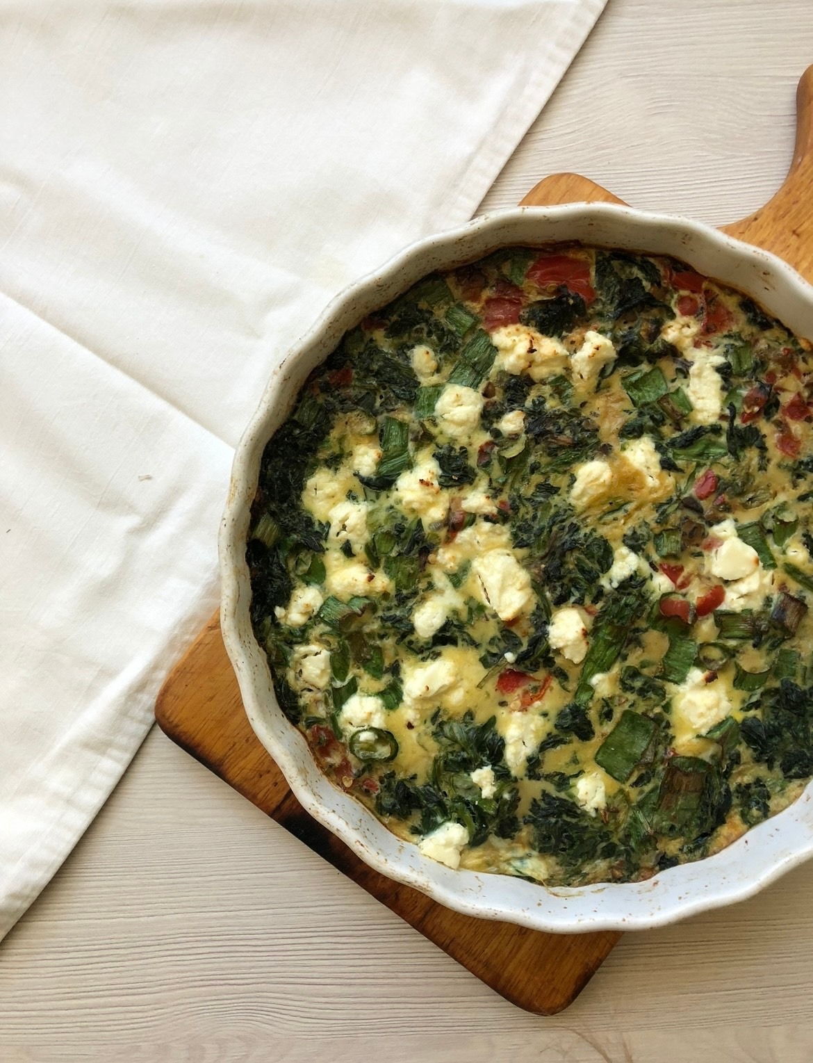 Quiche z zielonymi warzywami i serem feta w białej formie do pieczenia, ułożona na drewnianej desce, obok leży biała ściereczka na jasnym drewnianym stole, widok z góry.