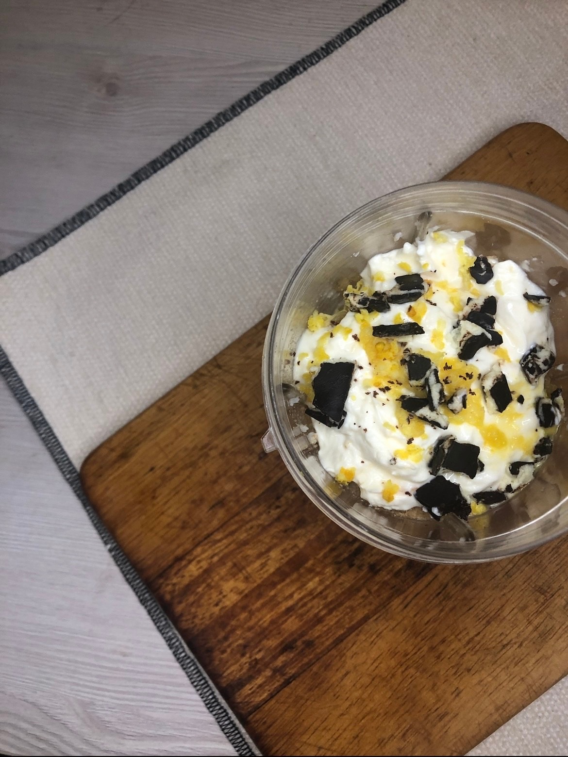 Deser w przezroczystym pojemniku: biały krem z pokruszonymi czarnymi ciastkami i skórką pomarańczową, na drewnianej desce obok szarej serwetki.