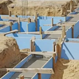 Szczegółowy widok wykopu z przygotowanymi szalunkami z niebieskiego styropianu i drewnianymi wzmocnieniami, częściowo wypełnionymi świeżym betonem na fundament domu.