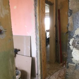 Julex Andrzej Cyganowki - Wnętrze w trakcie demontażu: odsłonięte mury, fragmenty tynku, widoczne przewody elektryczne, płyty gipsowo-kartonowe oparte o ścianę, poziomica oparta o ścianę w przejściu do innego pomieszczenia.