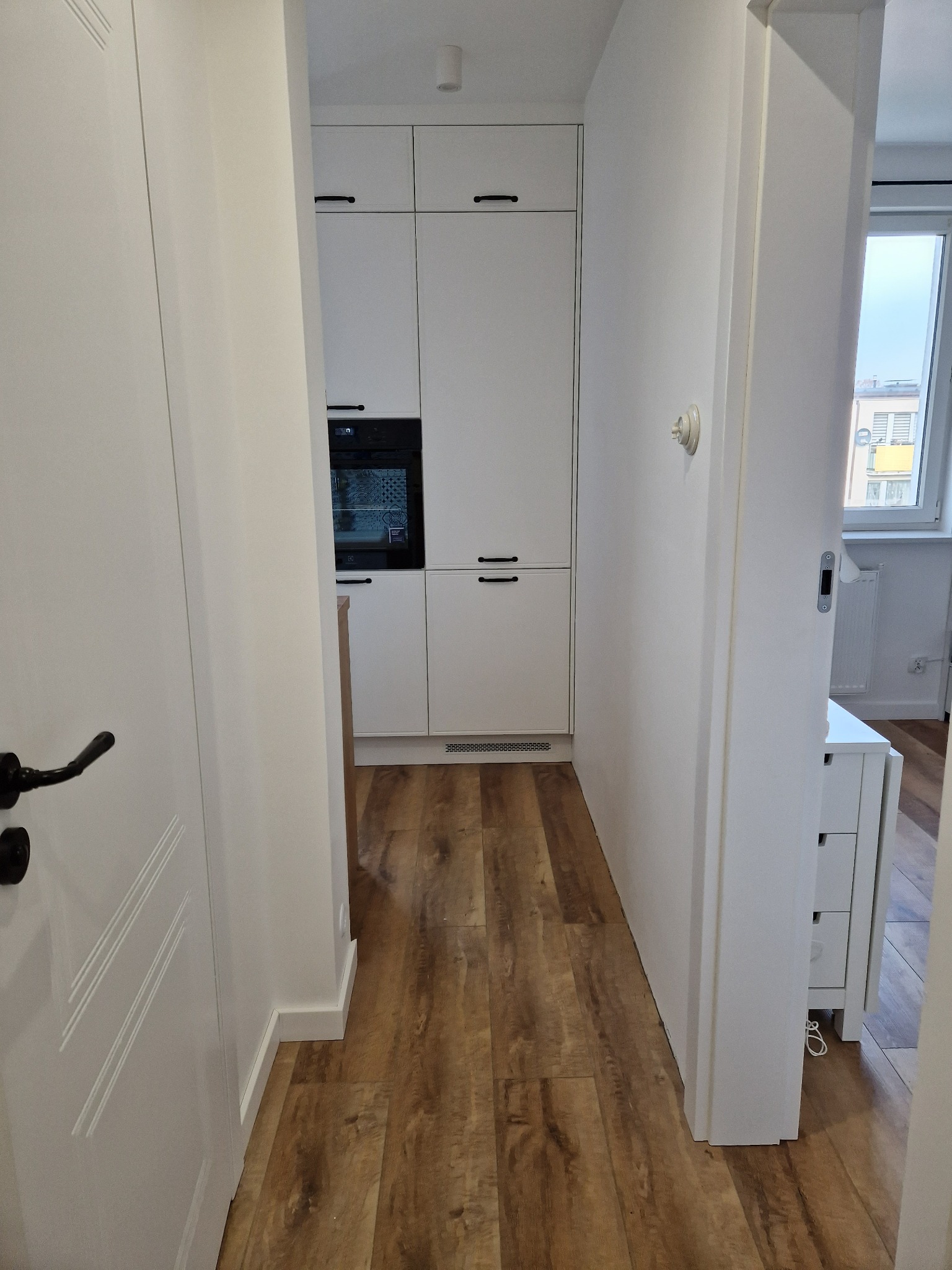 Wnętrze mieszkania z drewnianą podłogą i białą zabudową kuchenną z czarnymi uchwytami, widok z przedpokoju przez otwarte drzwi.