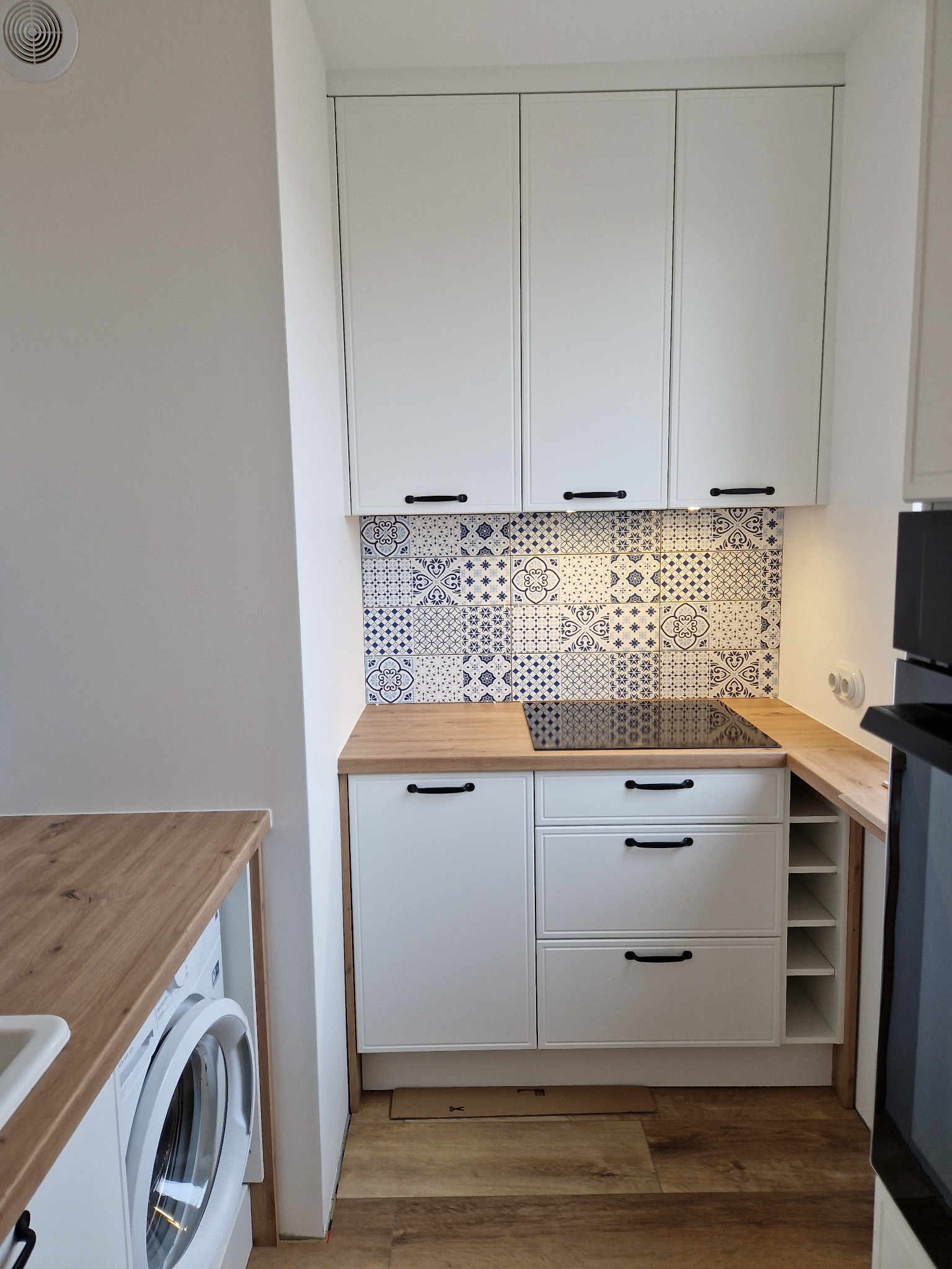 Nowoczesna, jasna kuchnia z białymi szafkami, drewnianym blatem i dekoracyjnymi, niebiesko-białymi płytkami nad indukcją; pralka w zabudowie po lewej stronie.