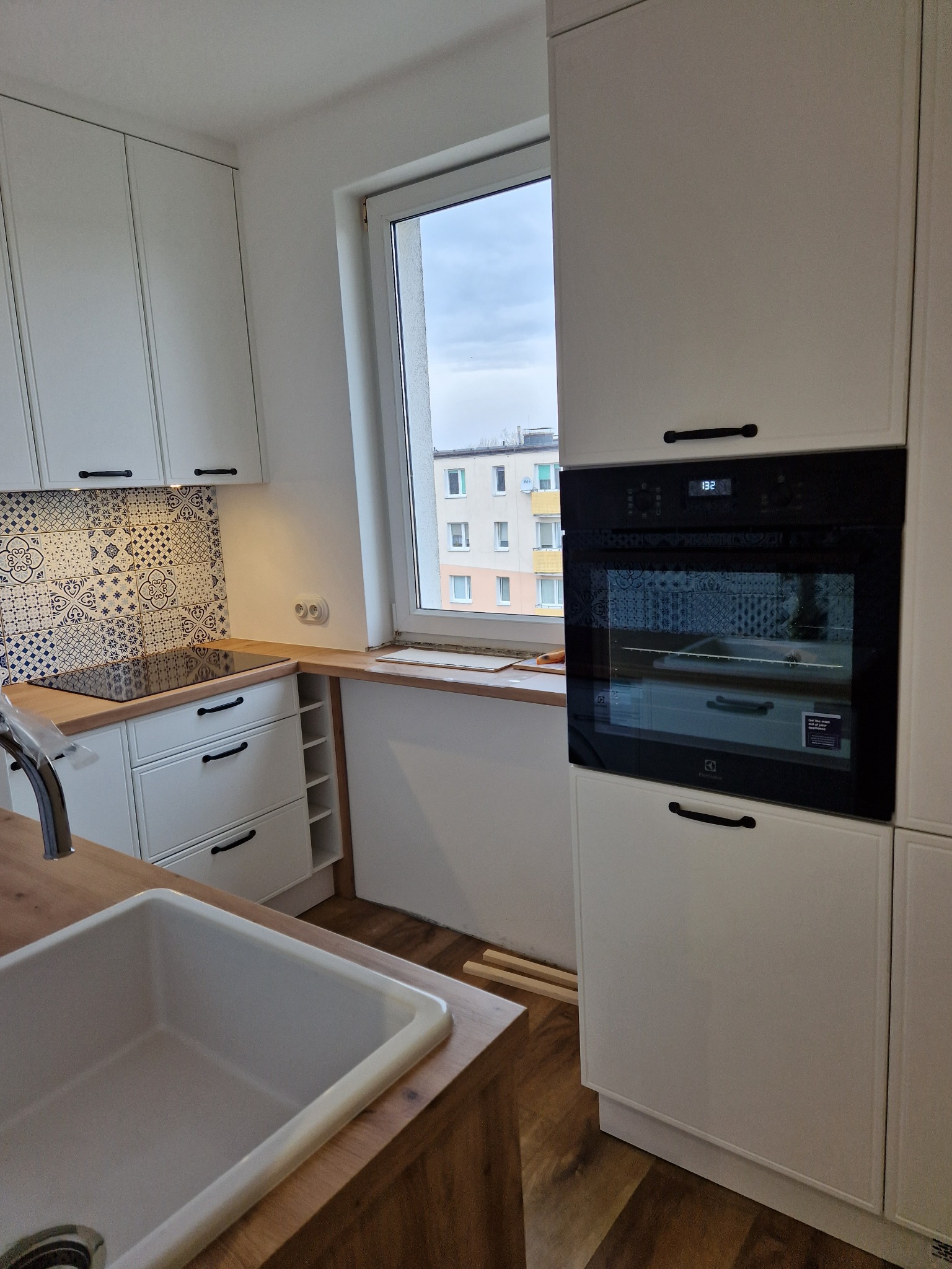 Nowoczesna kuchnia w trakcie wykańczania: białe szafki z czarnymi uchwytami, ceramiczna mozaika nad blatem, wbudowany piekarnik Electrolux, drewniany blat i widok z okna na budynek mieszkalny.