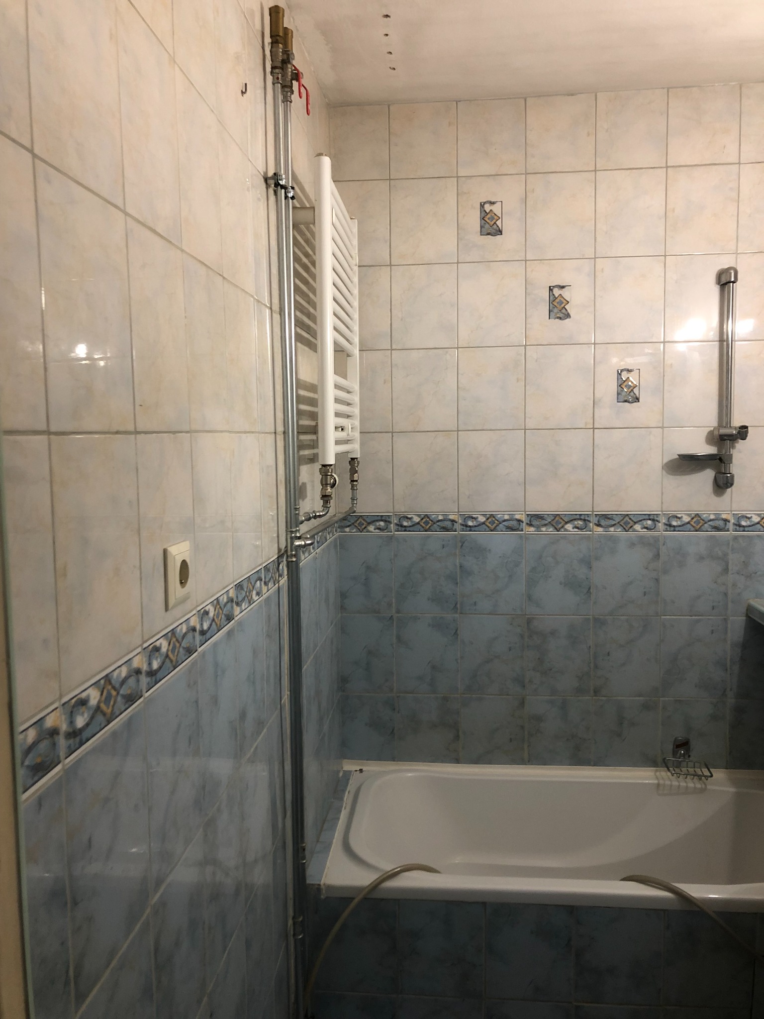 Biała drabinka grzejnikowa zamontowana na ścianie z kremowo-niebieskich płytek w łazience, widoczne rury doprowadzające wodę i fragment wanny.