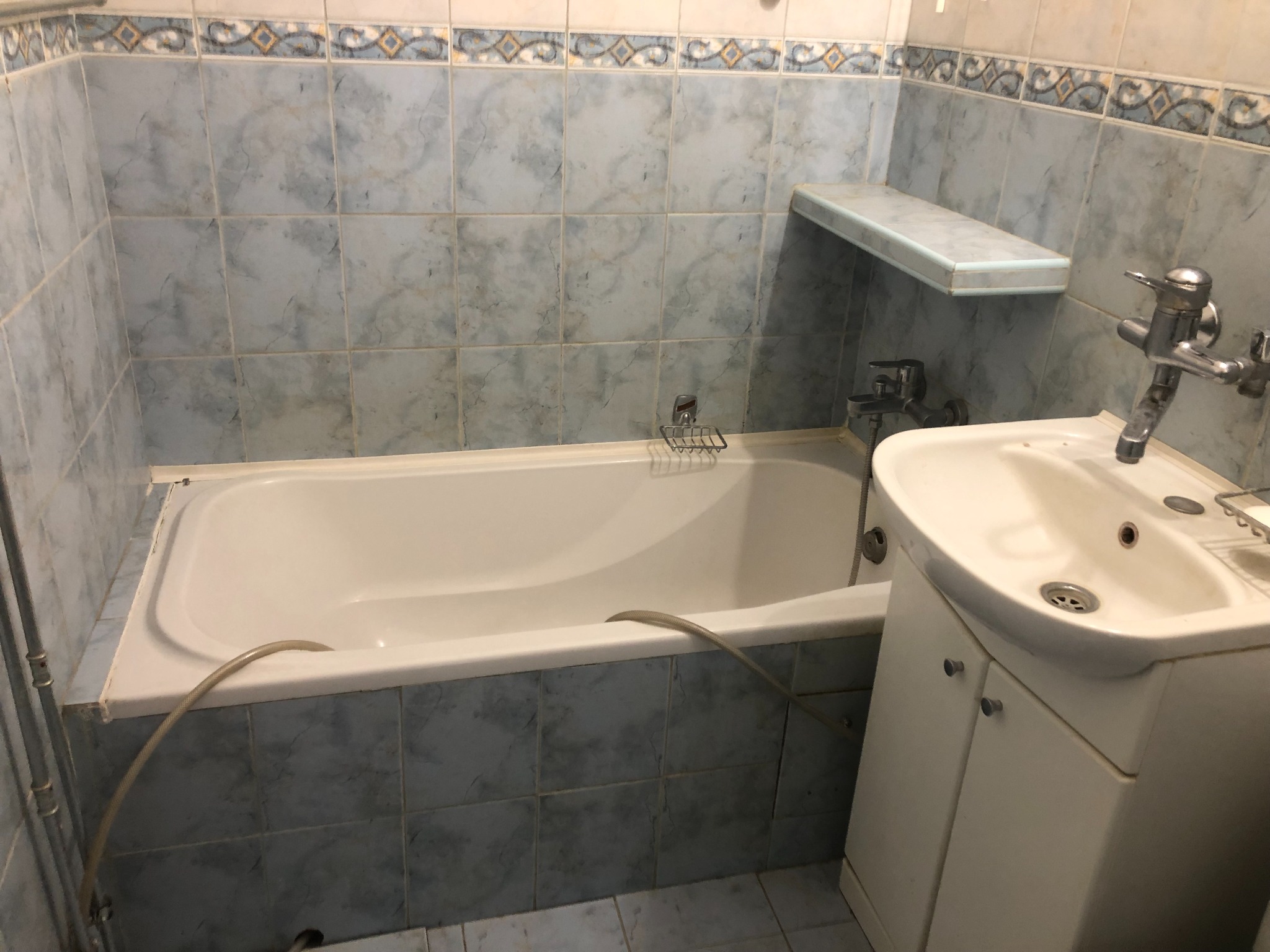 Standardowa łazienka z wanną obudowaną niebieskimi płytkami, umywalką z szafką i półką na ścianie, widoczne ślady użytkowania i wężyk w wannie.