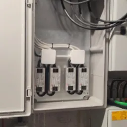 Otwarta skrzynka elektryczna z czterema modułami zabezpieczeń, widoczne przewody i złącze z czerwoną wtyczką na ścianie o surowym wykończeniu.