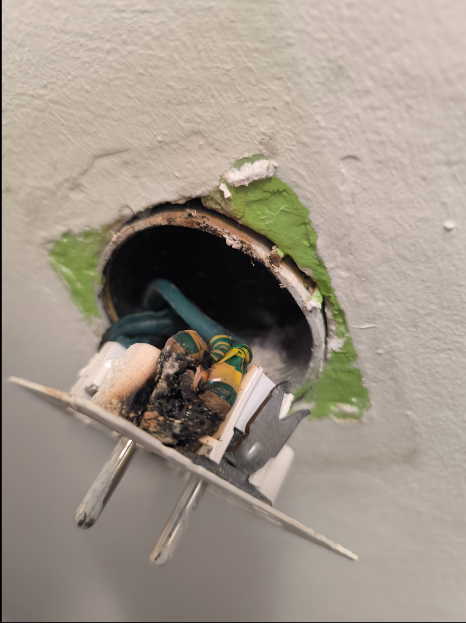 Uszkodzone gniazdko elektryczne wystające ze ściany z widocznymi zwęglonymi elementami i przewodami w izolacji, otoczone nierównym otworem w pomalowanej ścianie.