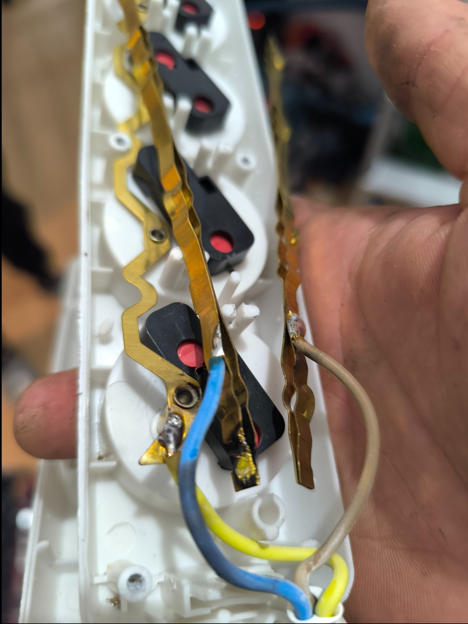 Detal przyłącza elektrycznego w gnieździe z widocznymi przewodami: niebieskim, żółtym i brązowym, lutowanymi do metalowych styków, trzymanego w dłoni.