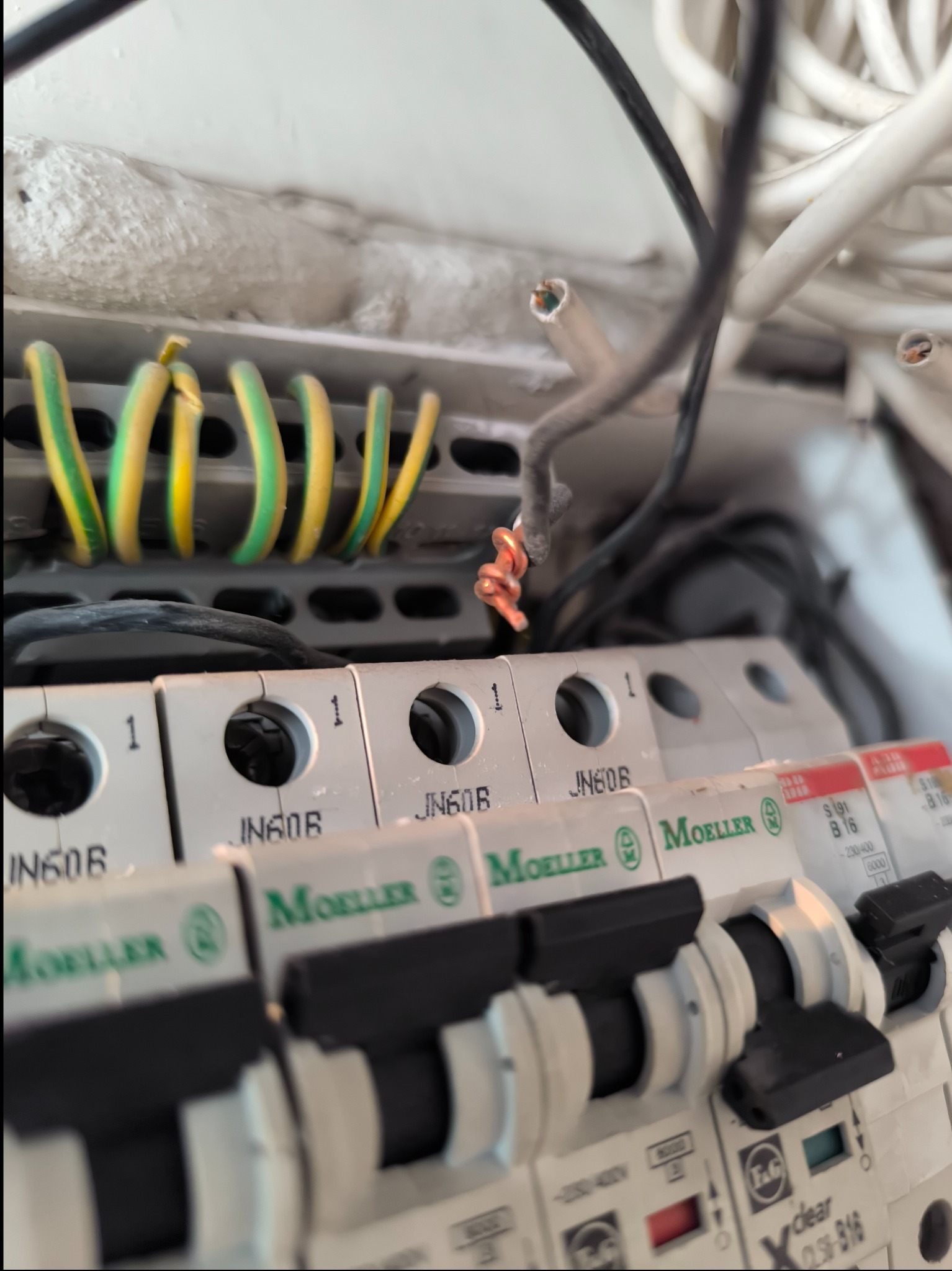 Otwarta skrzynka elektryczna z widocznymi bezpiecznikami Moeller JN60B, przewodami w izolacji żółto-zielonej oraz wystającymi, niezabezpieczonymi końcówkami miedzianych drutów.