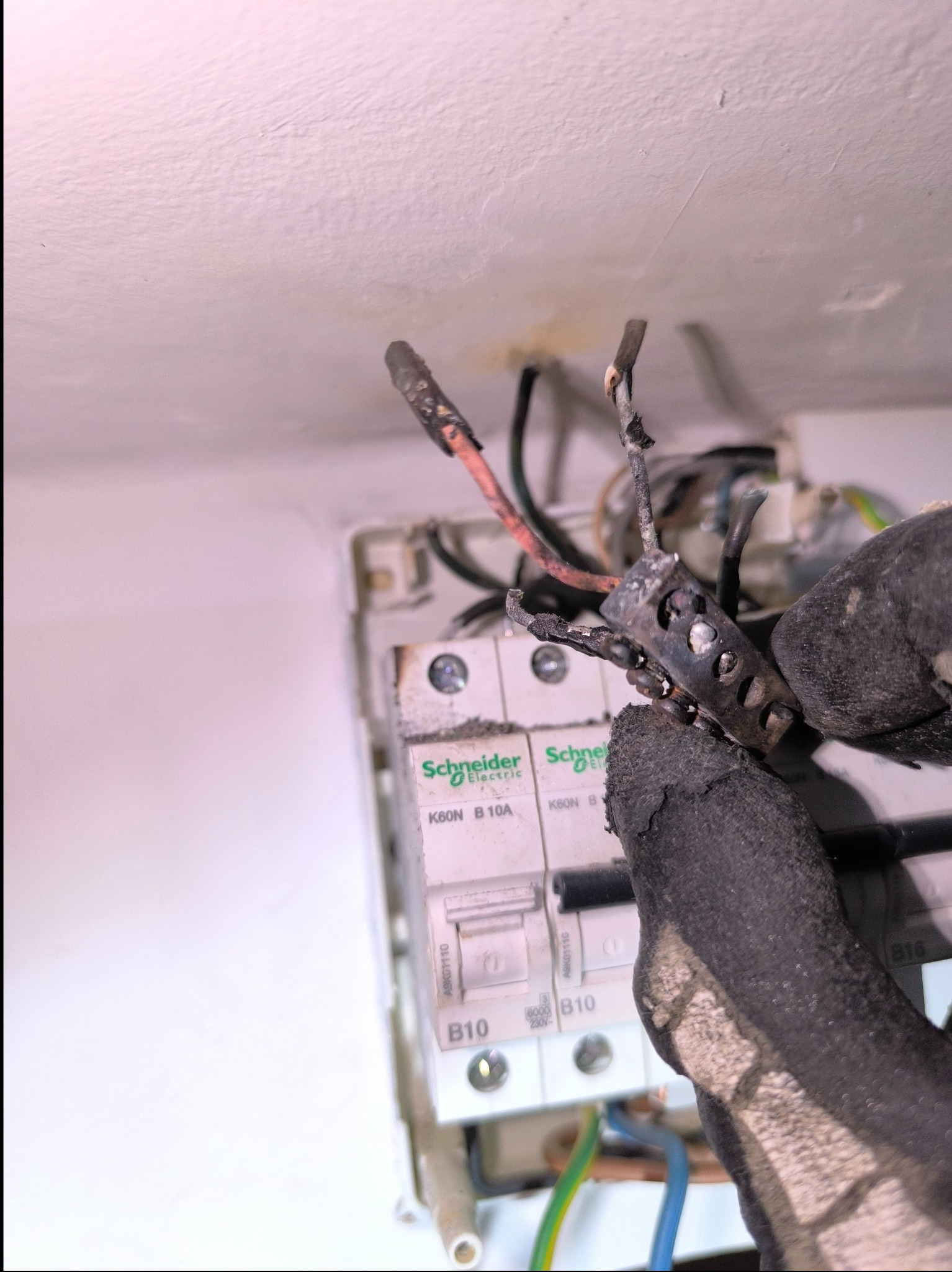 Przepalona kostka elektryczna w rękawicy roboczej na tle skrzynki z bezpiecznikami Schneider Electric, widoczne stopione elementy i uszkodzone przewody.