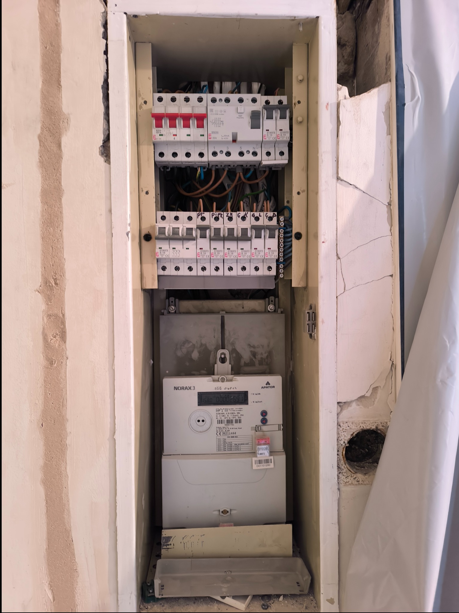 Tablica rozdzielcza z bezpiecznikami i licznikiem energii elektrycznej w obudowie w trakcie remontu, widoczne uszkodzenia ściany.