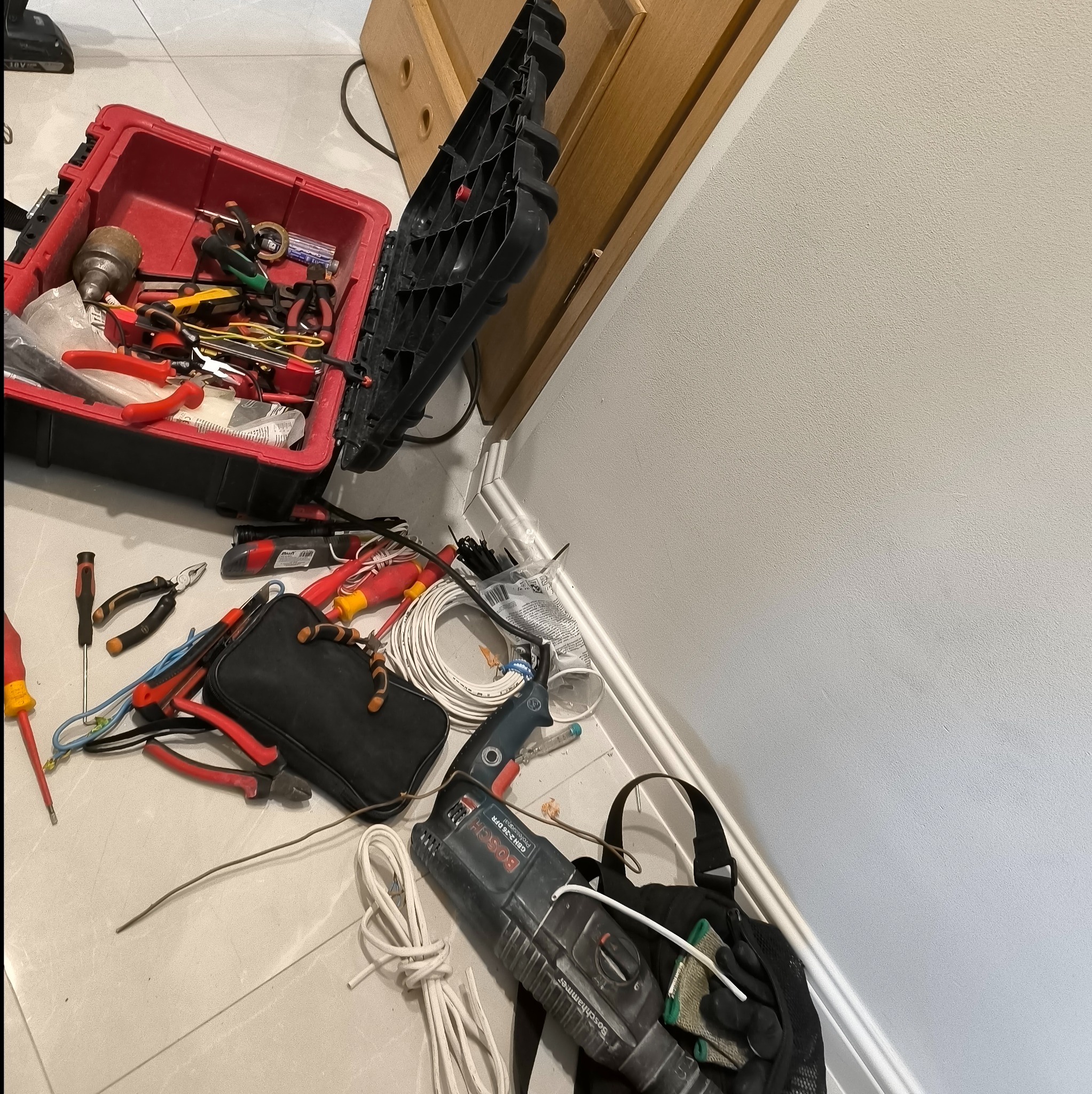 Rozłożona skrzynka z narzędziami elektrycznymi, w tym kombinerki, wkrętaki, przewody i wiertarka udarowa Bosch na podłodze z płytek, obok ściany i otwartych drzwi szafki.