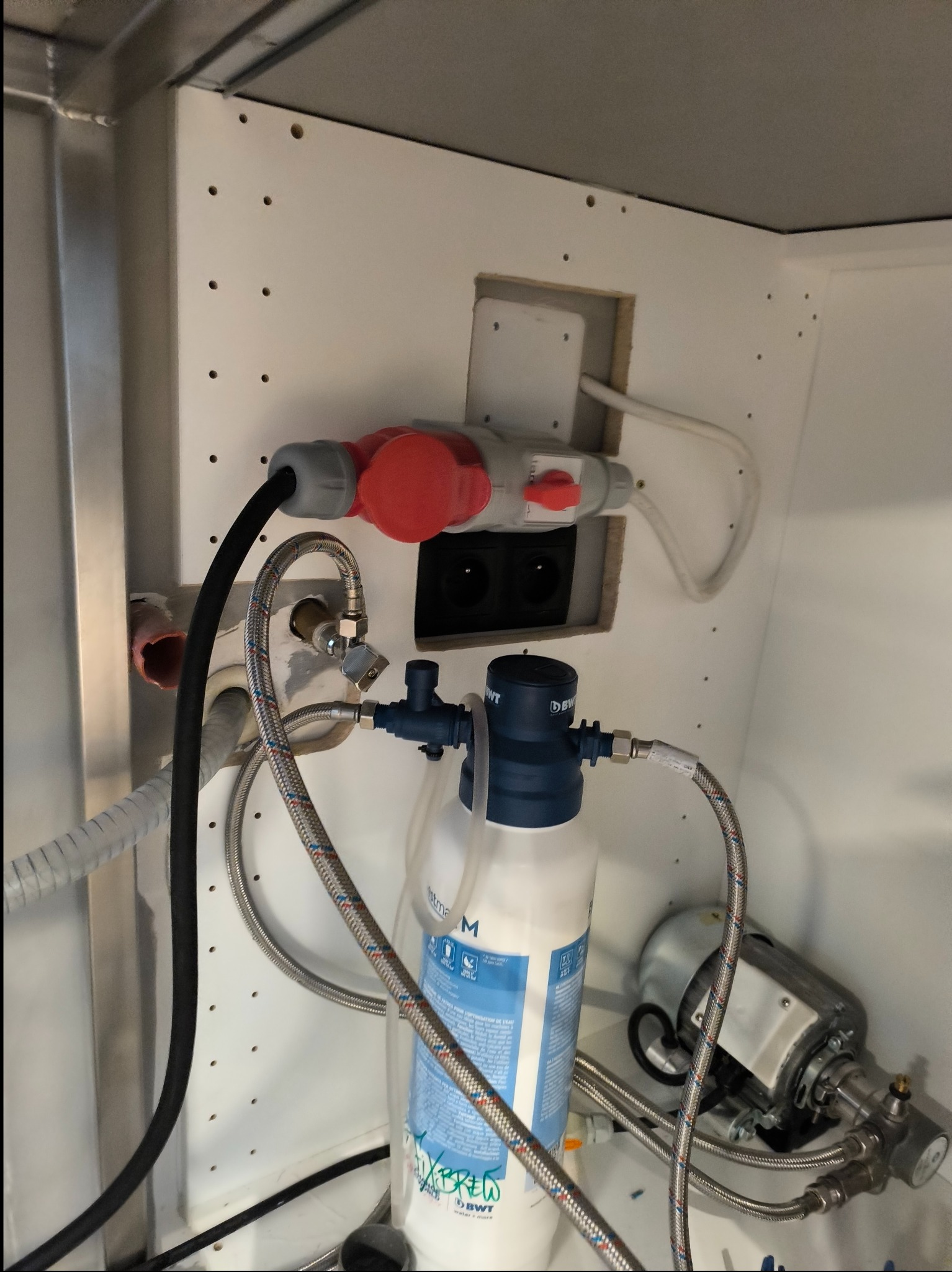 Częściowe ujęcie szafki z widoczną instalacją elektryczną (gniazdo siłowe) oraz elementami instalacji wodnej: filtr wody BWT, metalowe wężyki i pompa.