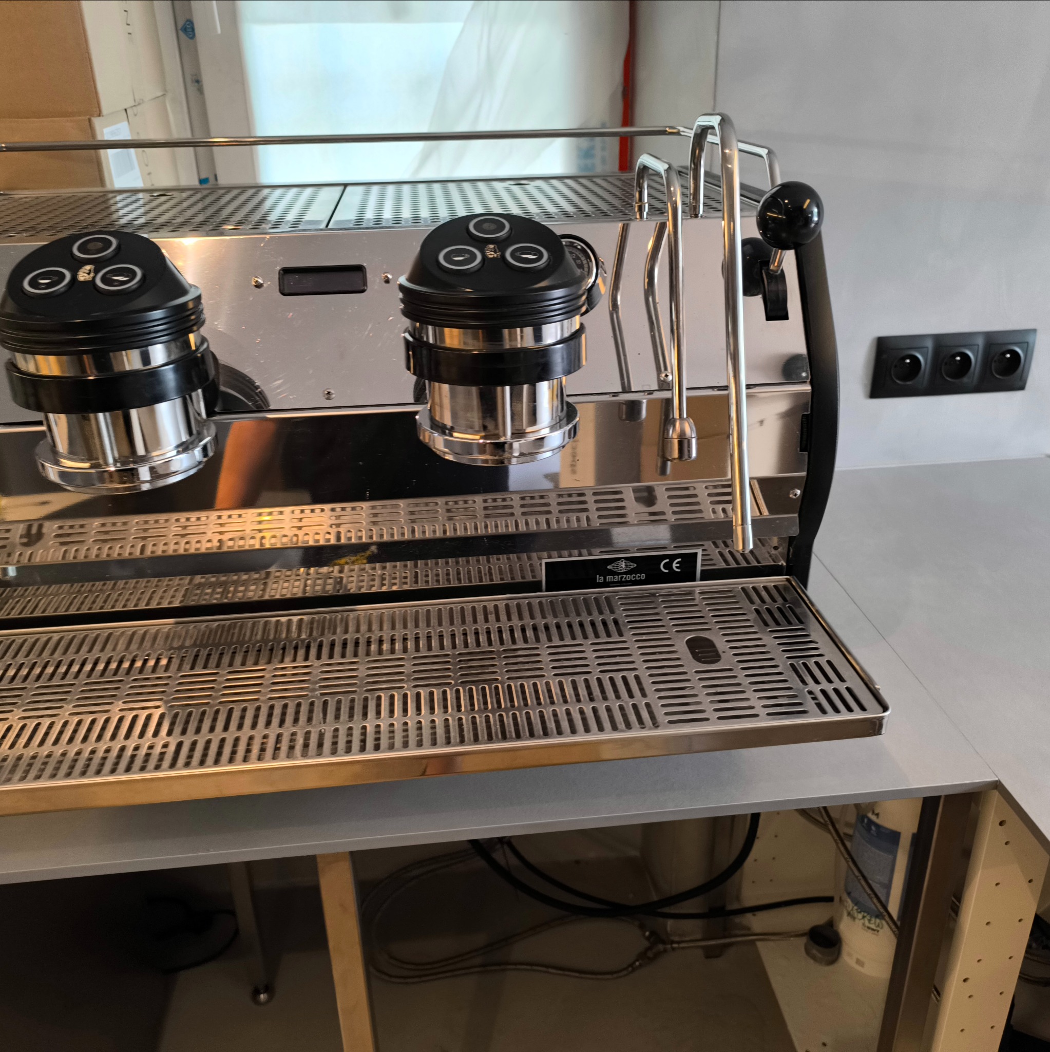 Profesjonalny ekspres do kawy La Marzocco ze stali nierdzewnej, widoczne przyłącza elektryczne i hydrauliczne pod blatem.