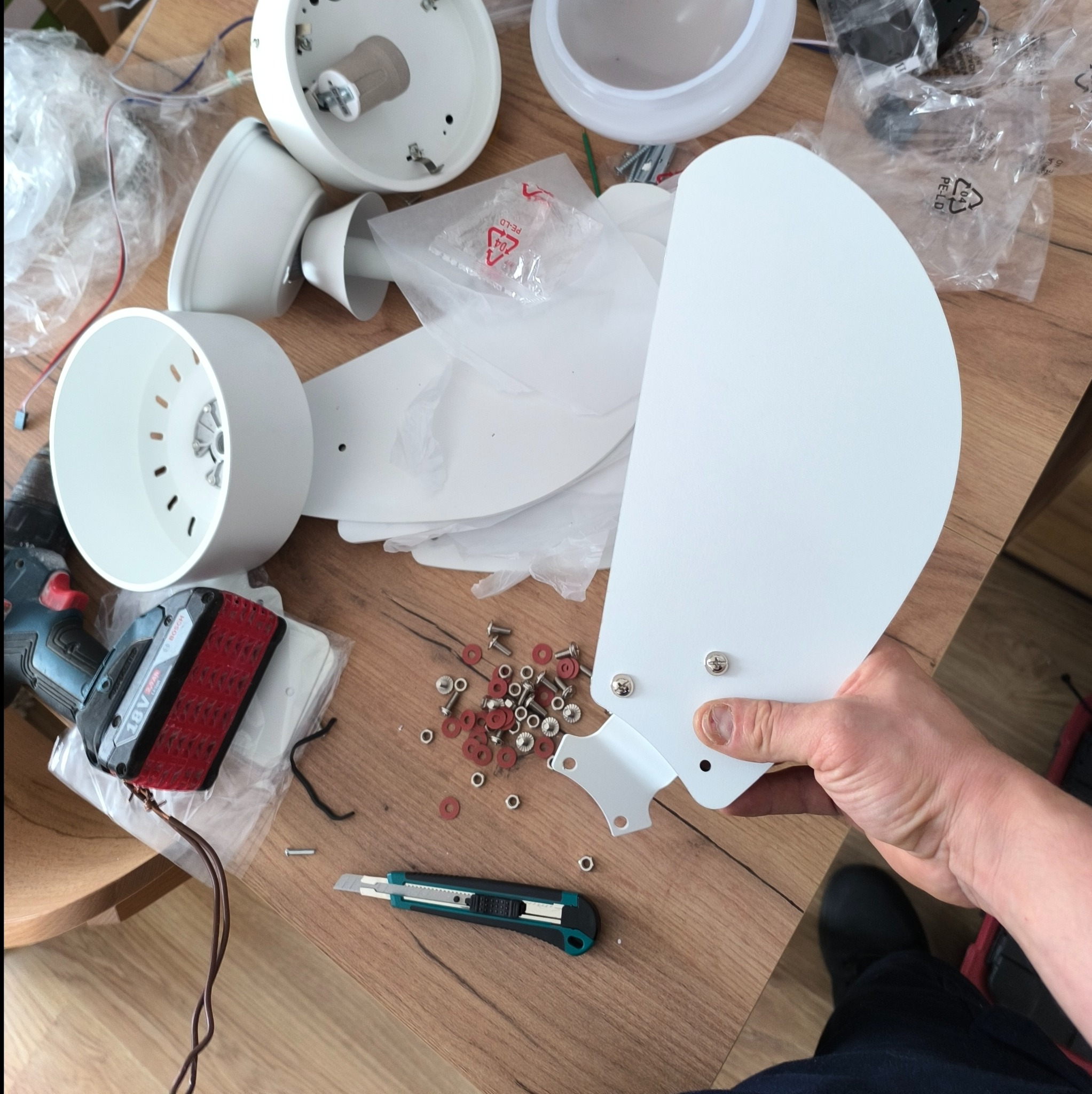 Elementy składowe białego wentylatora sufitowego rozłożone na drewnianym stole, w tym obudowa, łopatki, śrubki, wkrętarka Bosch i nóż monterski.