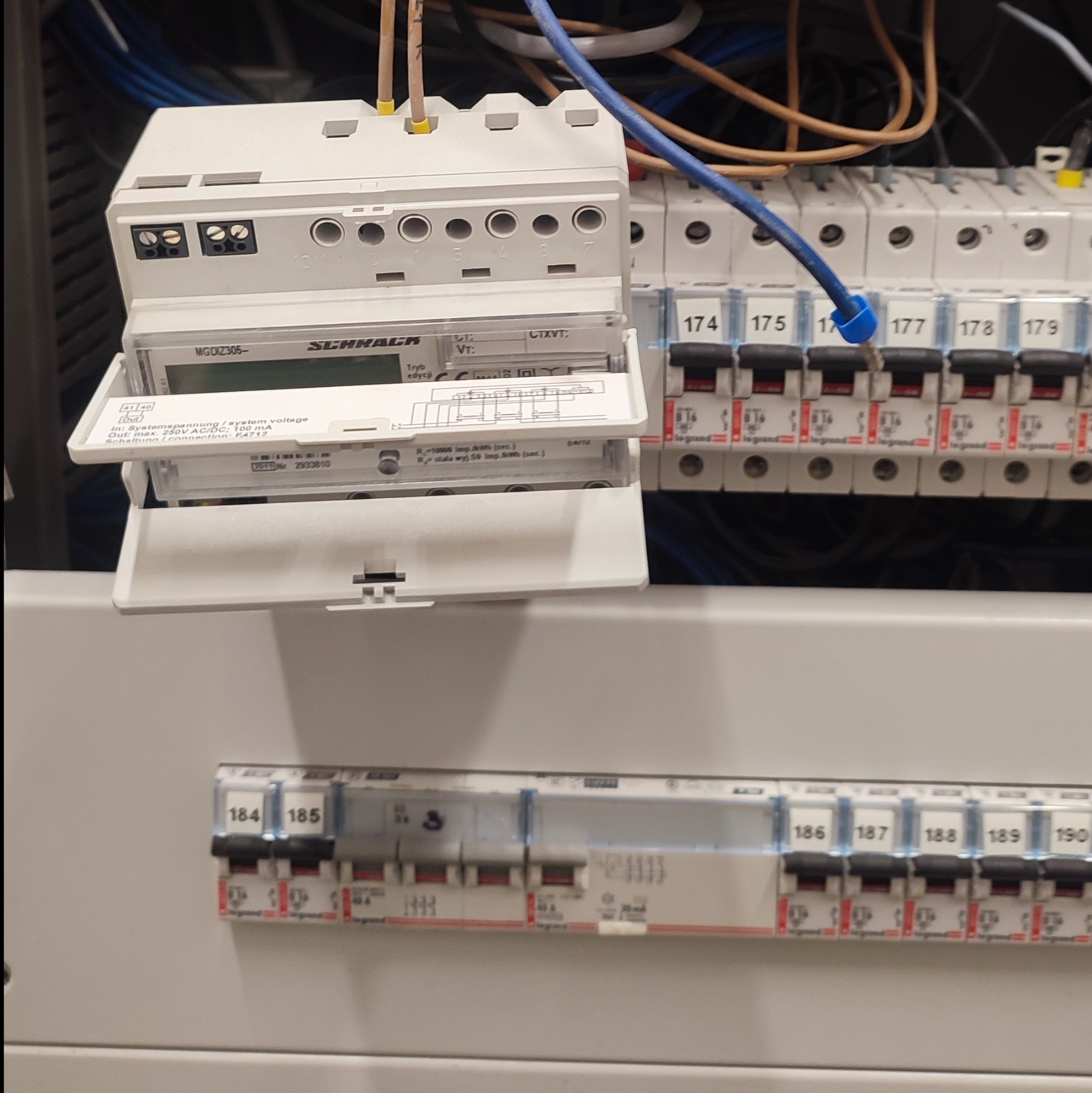 Szczegółowe ujęcie tablicy rozdzielczej z zamontowanym licznikiem energii elektrycznej Schrack, widoczne bezpieczniki z numeracją 174-190 oraz podłączone przewody.
