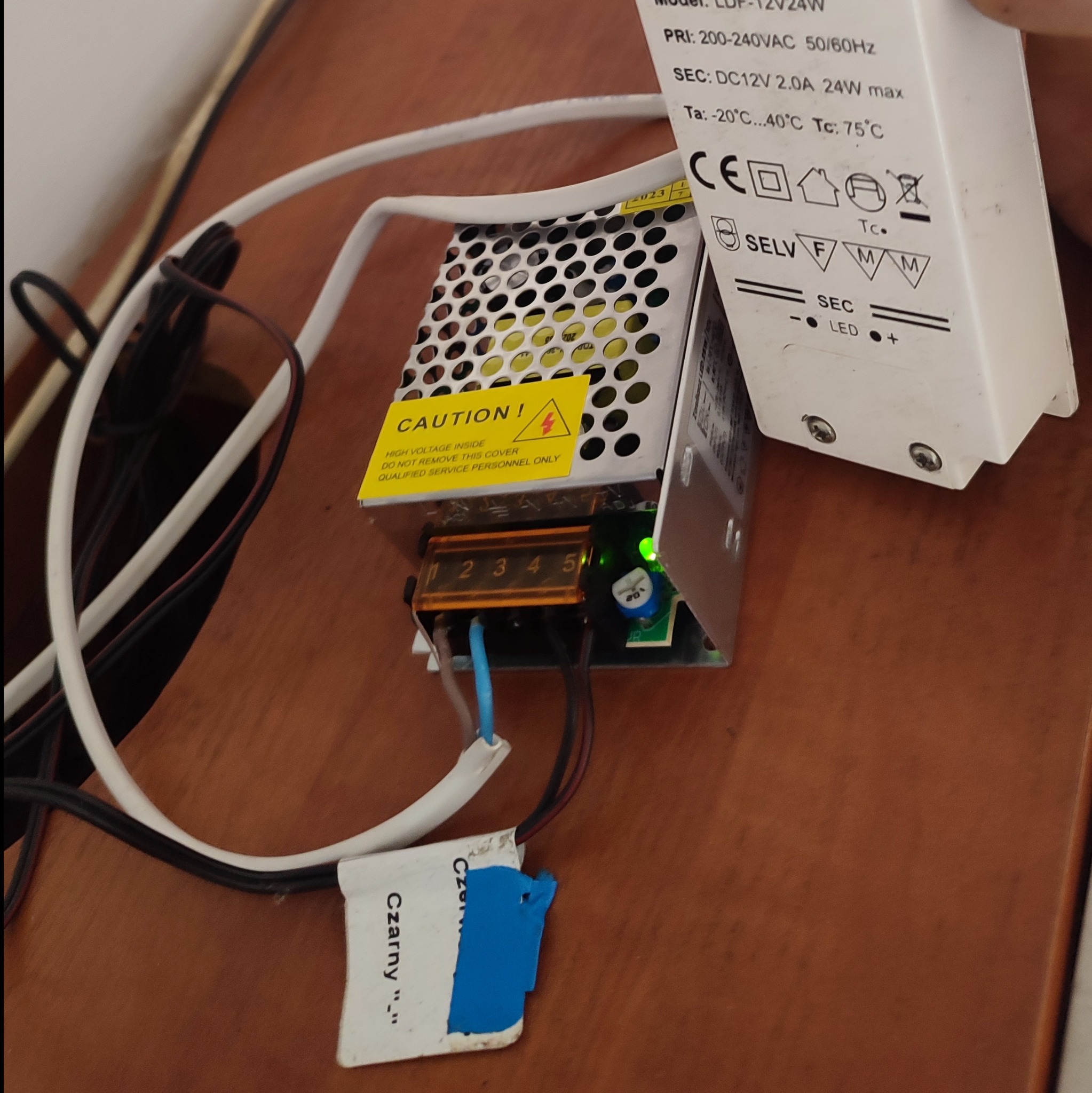 Zasilacz LED 12V24W z widocznymi przewodami, kostką podłączeniową, naklejką ostrzegawczą i specyfikacją techniczną, leżący na drewnianej powierzchni.