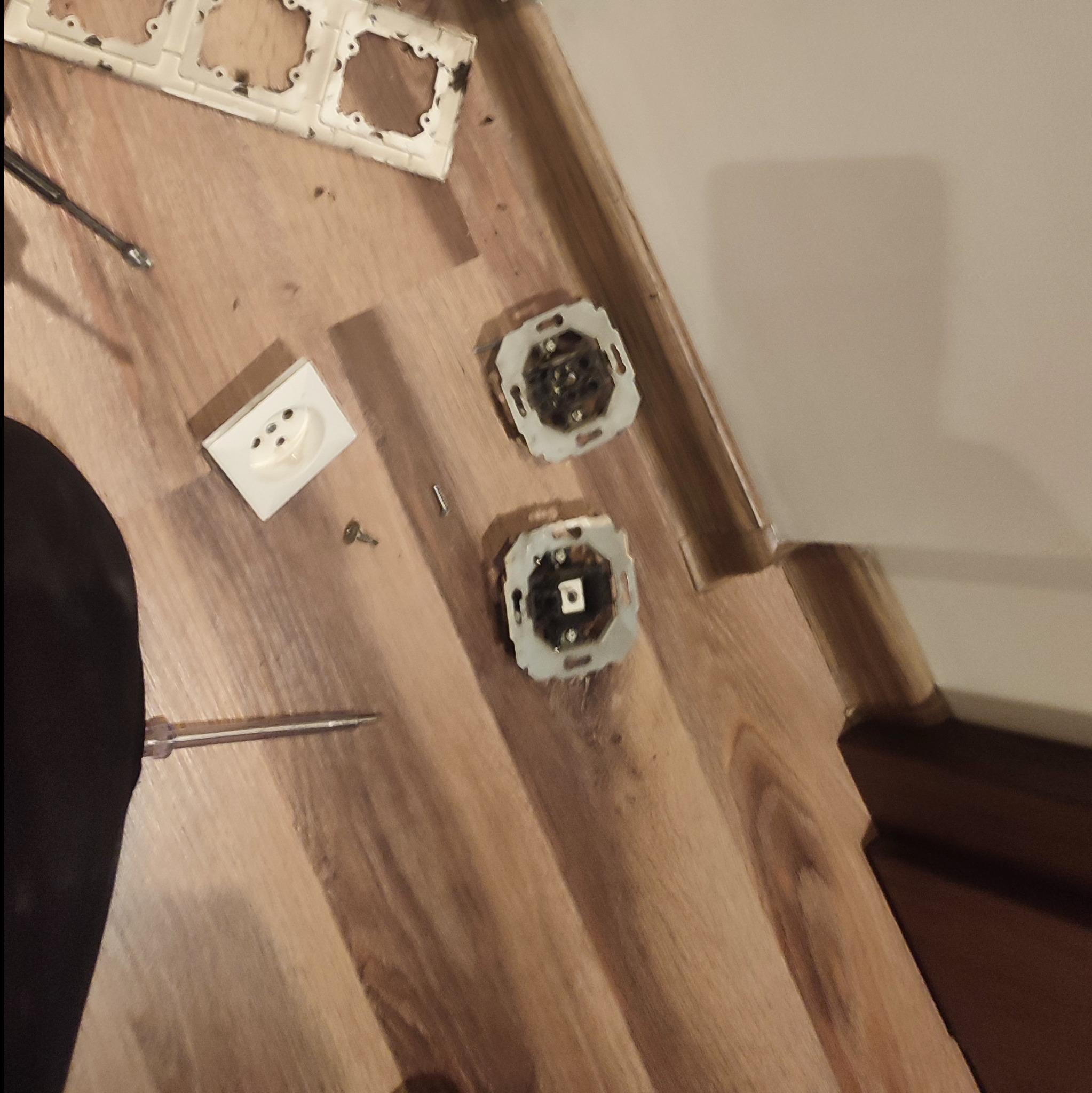 Demontaż gniazdek elektrycznych i włączników na drewnianej podłodze, widoczne ślady użytkowania, śrubokręt i elementy mocujące obok.