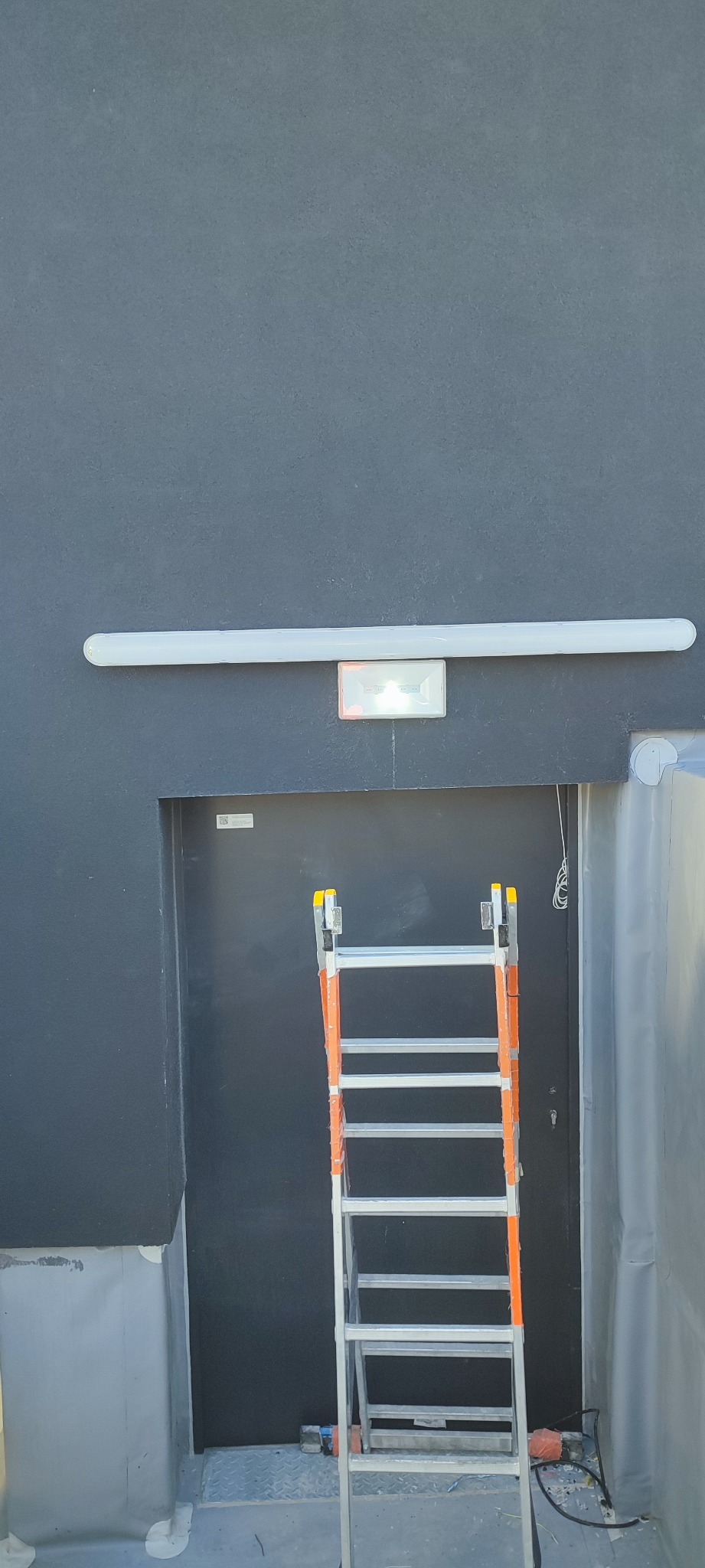 Zamontowana lampa jarzeniowa i lampa awaryjna nad wejściem, widoczna drabina aluminiowa i narzędzia podczas prac instalacyjnych.