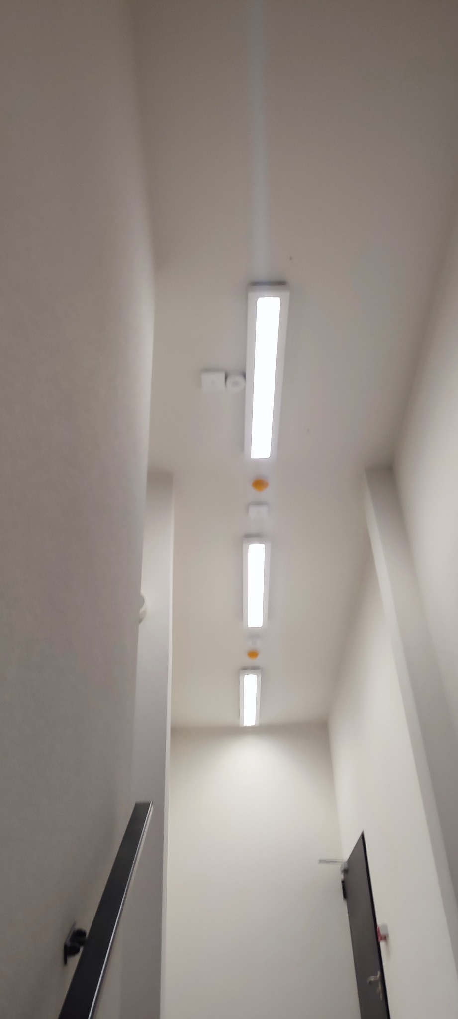 Wnętrze klatki schodowej z trzema podłużnymi lampami LED zamontowanymi na suficie, czujnikiem dymu i drzwiami na końcu korytarza.