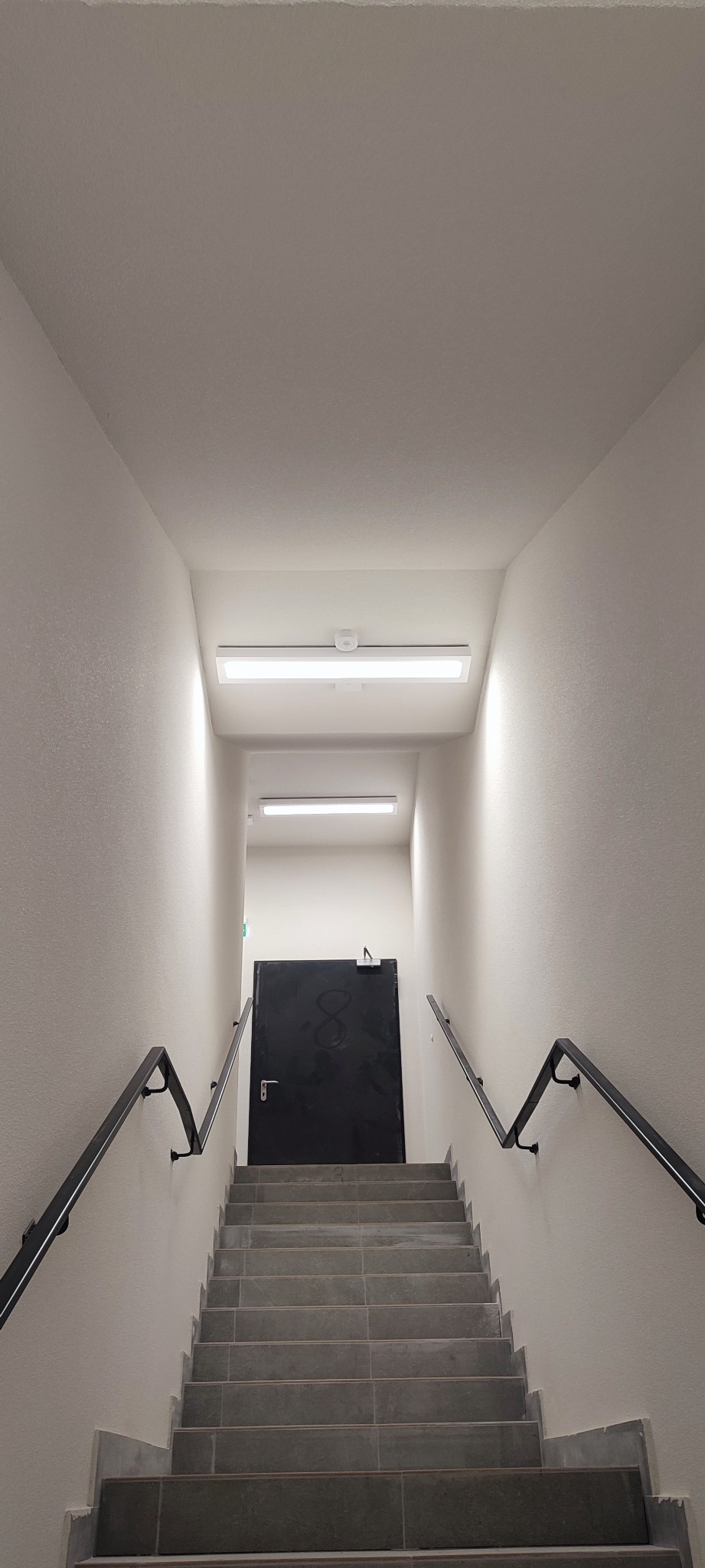 Klatka schodowa z szarymi płytkami, czarną poręczą i drzwiami z numerem 8 w tle, oświetlona lampami LED.