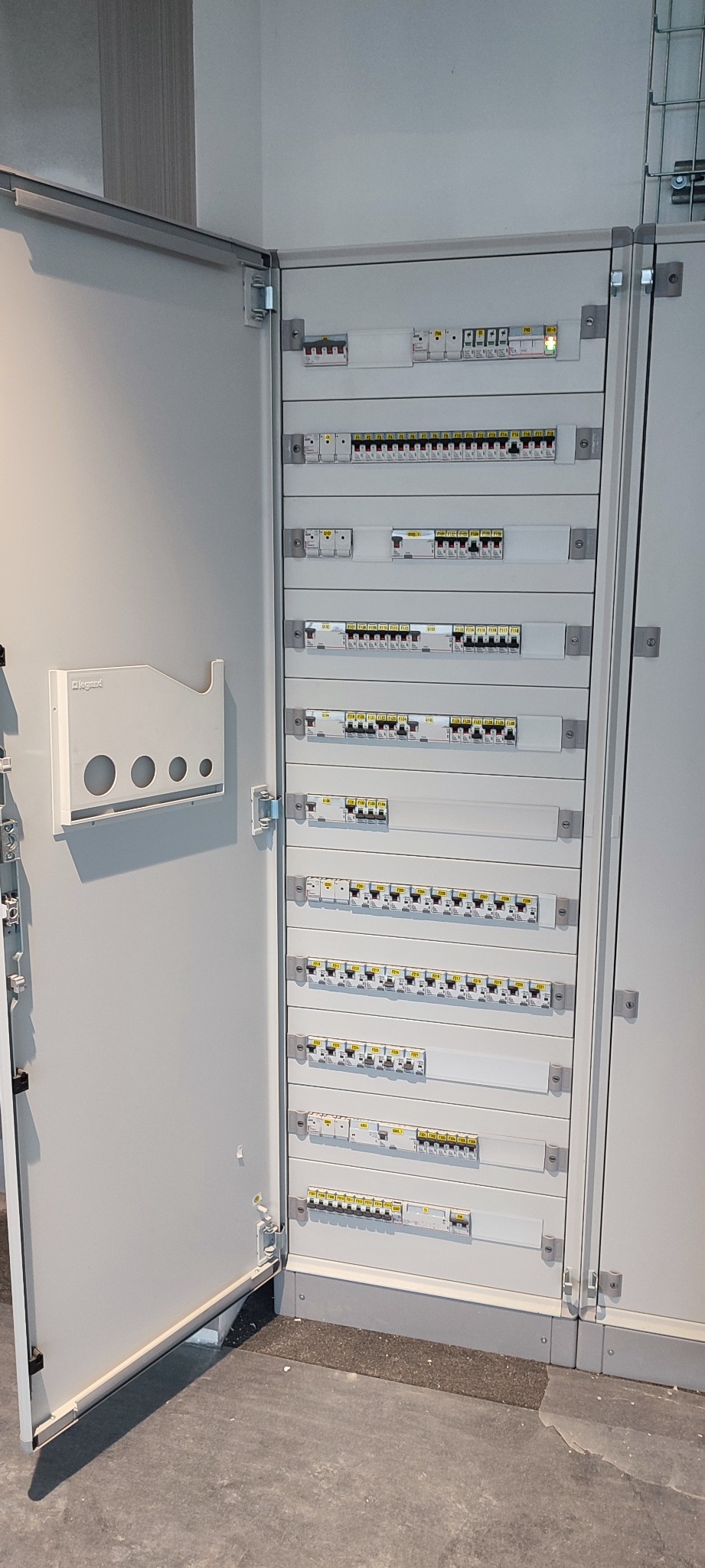 Otwarta skrzynka elektryczna z wieloma rzędami bezpieczników i wyłączników, widoczna półka na dokumenty po lewej stronie.