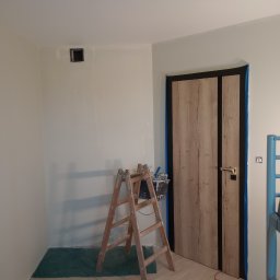 Grzegorz Kowalski - Pokój w trakcie odświeżania: drabina malarska z wiadrem farby stoi przy ścianie z zamaskowanym otworem wentylacyjnym, drzwi oklejone taśmą malarską, podłoga częściowo zakryta folią ochronną.