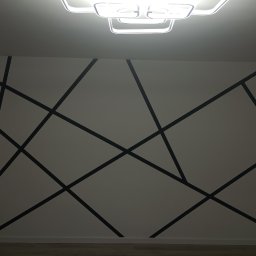 Grzegorz Kowalski - Ściana pomalowana w geometryczny wzór z czarnych linii na białym tle, oświetlona nowoczesną lampą sufitową.