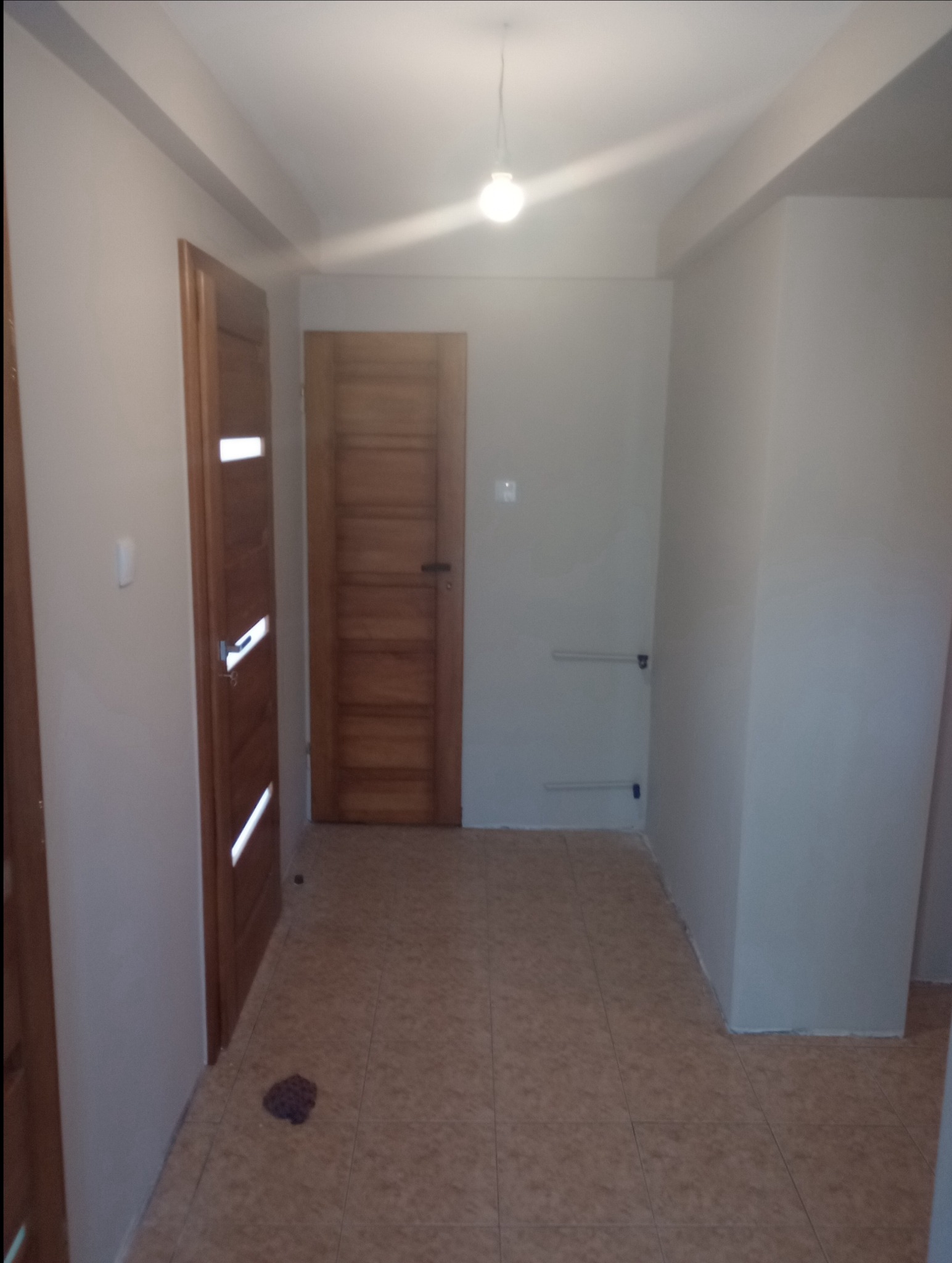 Wnętrze korytarza po malowaniu ścian na jasny kolor, z dwiema drewnianymi ościeżnicami drzwiowymi i brązową podłogą z płytek, widoczne rury instalacyjne przy ścianie.