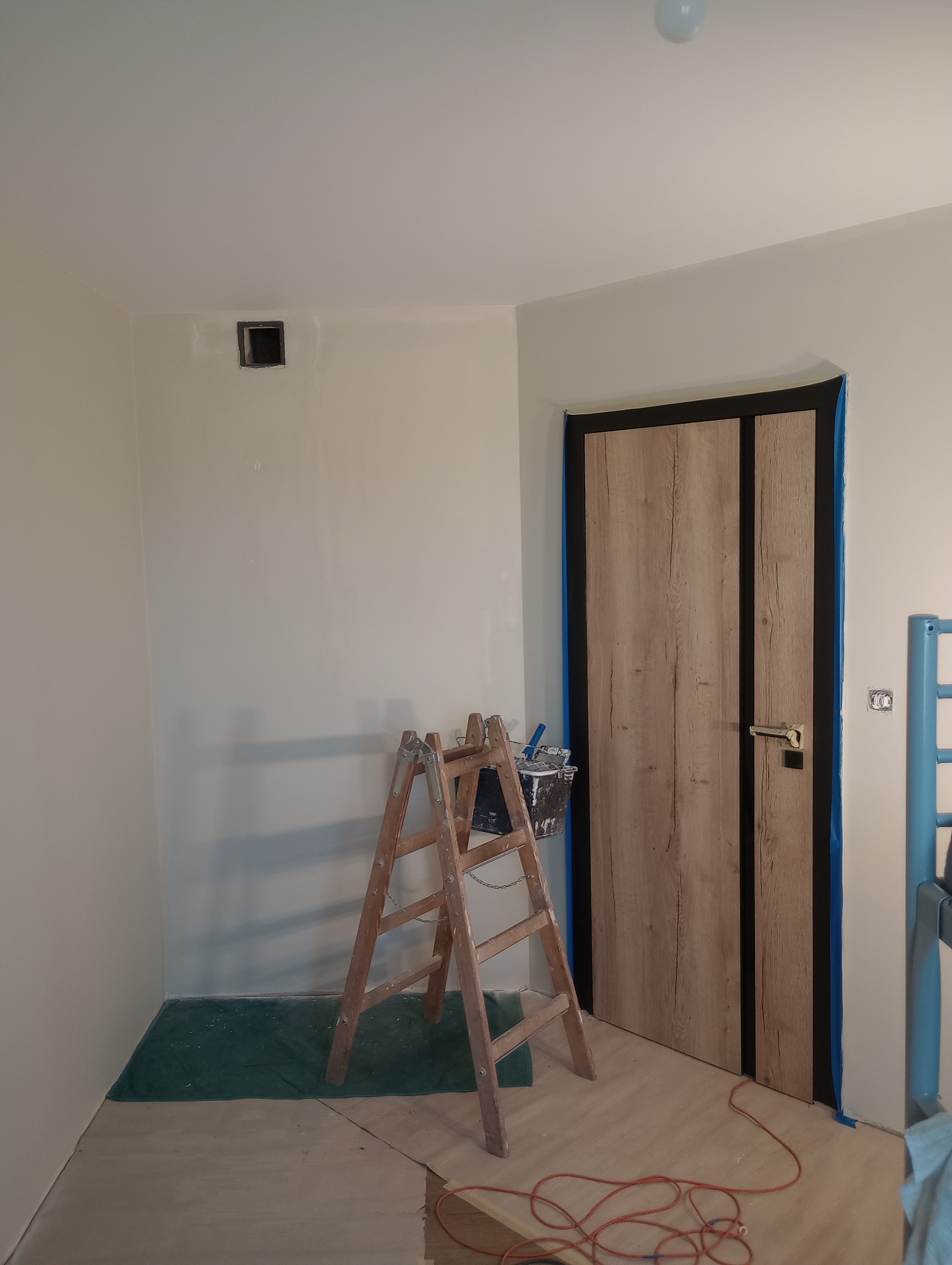 Pokój w trakcie odświeżania: drabina malarska z wiadrem farby stoi przy ścianie z zamaskowanym otworem wentylacyjnym, drzwi oklejone taśmą malarską, podłoga częściowo zakryta folią ochronną.