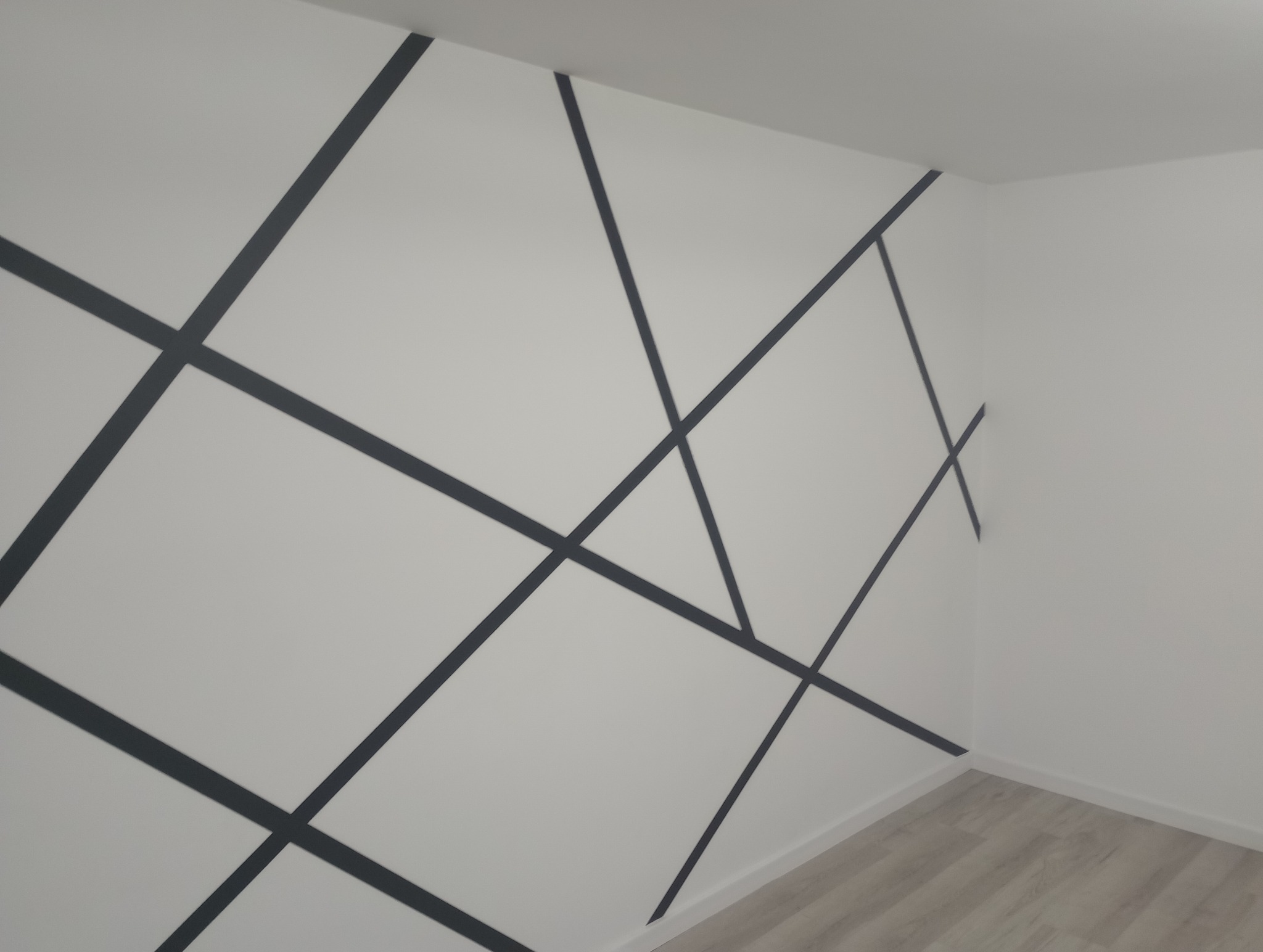 Wnętrze pokoju po malowaniu z geometrycznym wzorem z ciemnych pasów na białych ścianach, widoczny fragment podłogi z paneli.