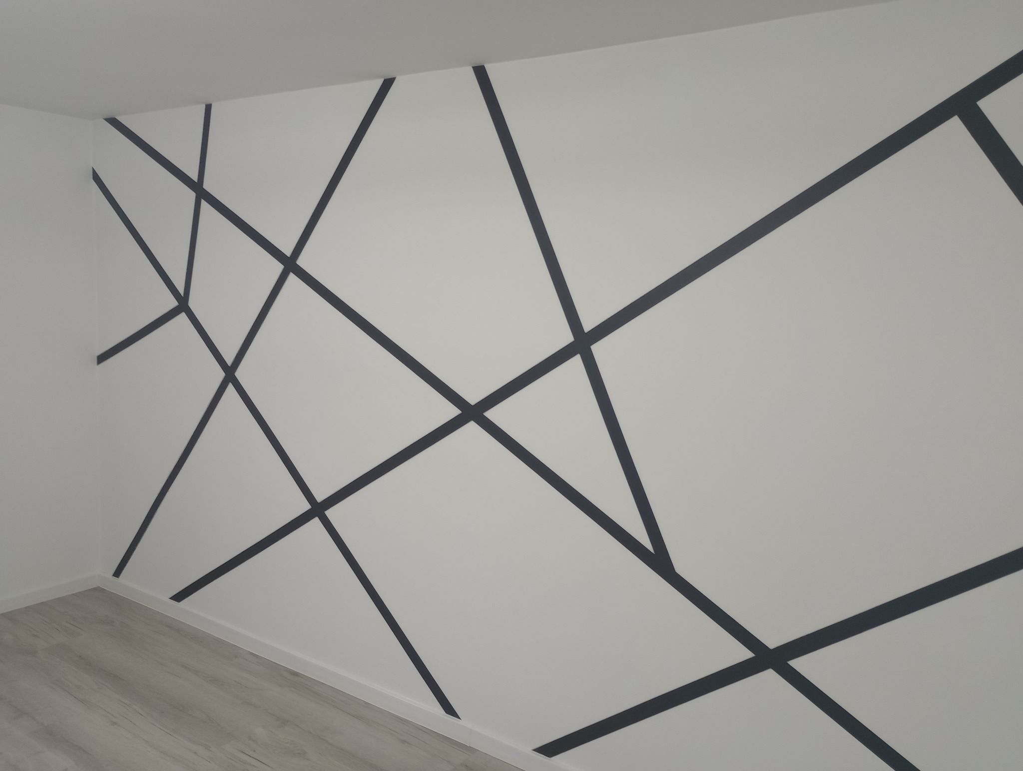 Biała ściana z geometrycznym wzorem z ciemnych linii, świeżo po realizacji w Lututowie, z widoczną podłogą z paneli.