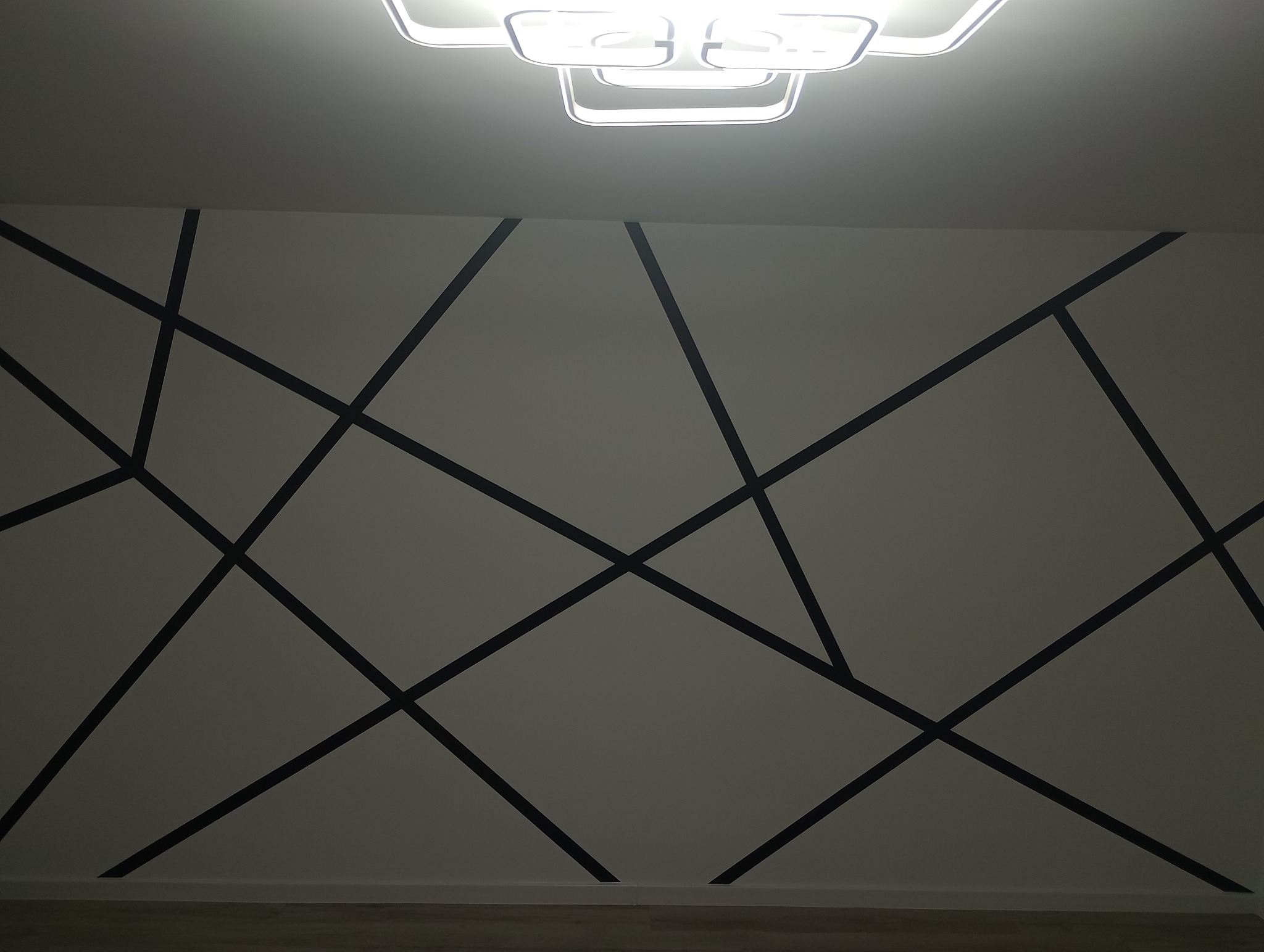 Ściana pomalowana w geometryczny wzór z czarnych linii na białym tle, oświetlona nowoczesną lampą sufitową.