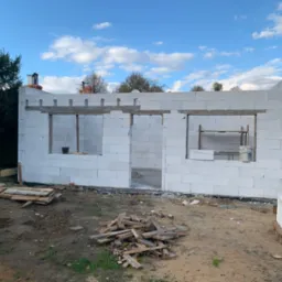 Wznoszenie ścian domu z białych bloczków, widoczne otwory okienne i drzwiowe, na ziemi porozrzucane drewniane elementy i paleta z bloczkami, w tle drzewa i niebo z chmurami.