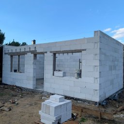 Tomasz kudela - Budowa domu z bloczków betonowych: ściany w trakcie wznoszenia, widoczne otwory okienne i drzwiowe z drewnianymi nadprożami, paleta z bloczkami i pies rasy buldog francuski w tle.