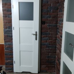 Tomasz kudela - Białe drzwi wewnętrzne z szybą, zamontowane w ścianie z cegły dekoracyjnej, widoczne fragmenty podłogi imitującej drewno oraz częściowo widoczne kolejne drzwi.