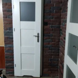 Białe drzwi wewnętrzne z szybą, zamontowane w ścianie z cegły dekoracyjnej, widoczne fragmenty podłogi imitującej drewno oraz częściowo widoczne kolejne drzwi.
