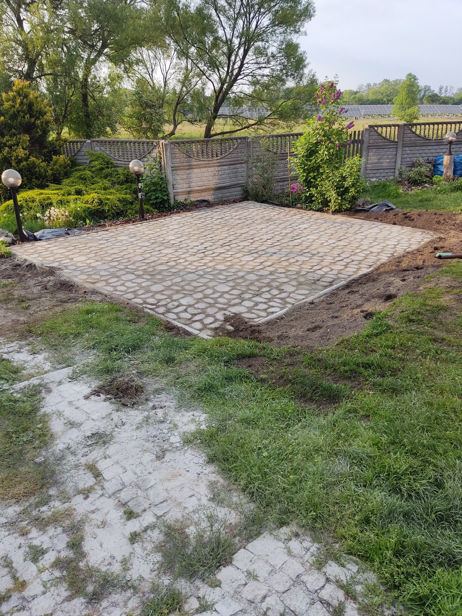 Przygotowany teren pod budowę tarasu z ułożoną kostką brukową o nieregularnym kształcie, otoczony świeżo przekopaną ziemią i trawnikiem, widoczne lampy ogrodowe i ozdobny płot w tle.