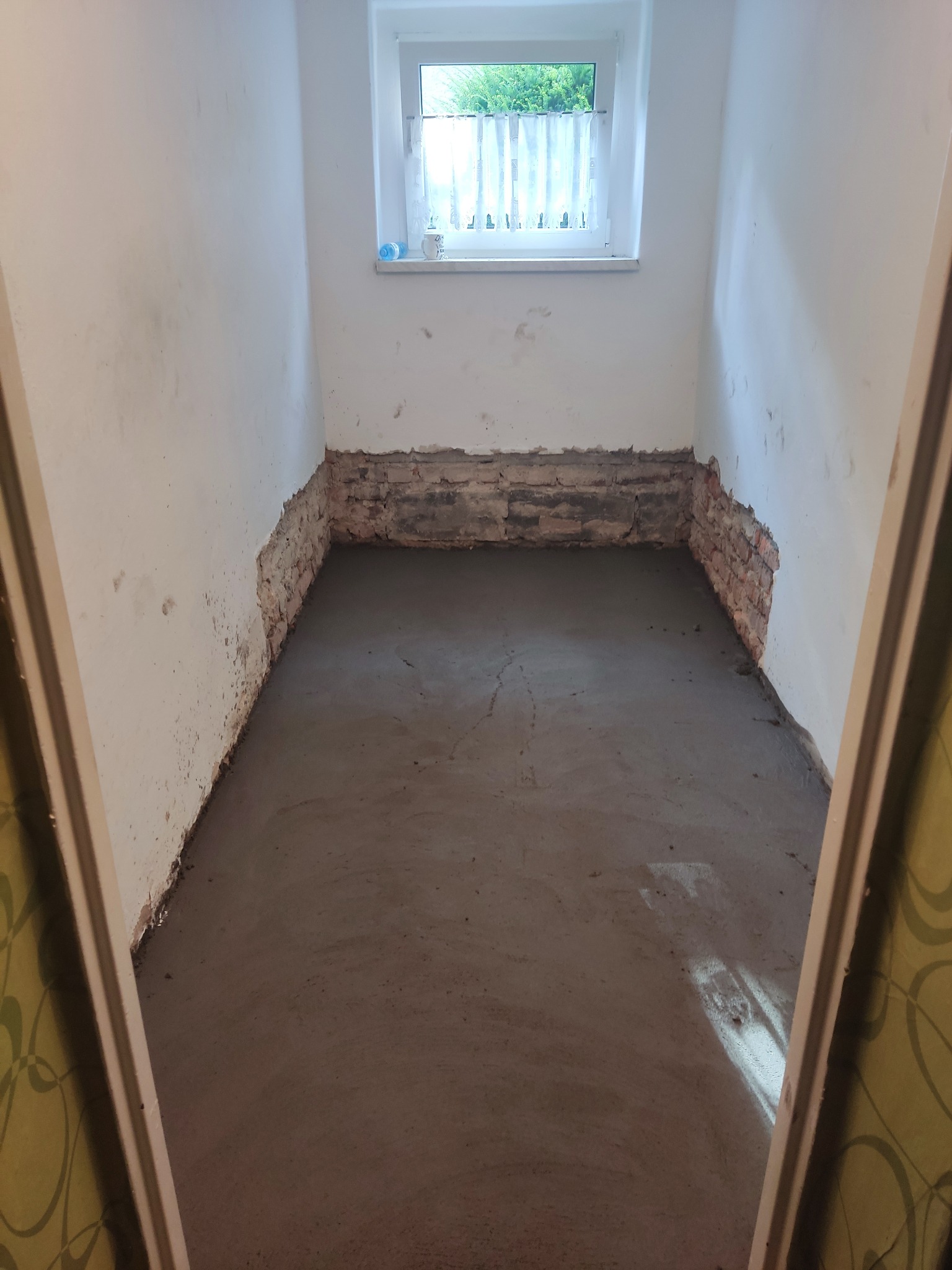 Wnętrze pomieszczenia w trakcie remontu, z wylewką betonową na podłodze i odsłoniętą cegłą u dołu ścian, widoczne przez framugę drzwi z wzorzystą tapetą i małe okno z firanką w tle.