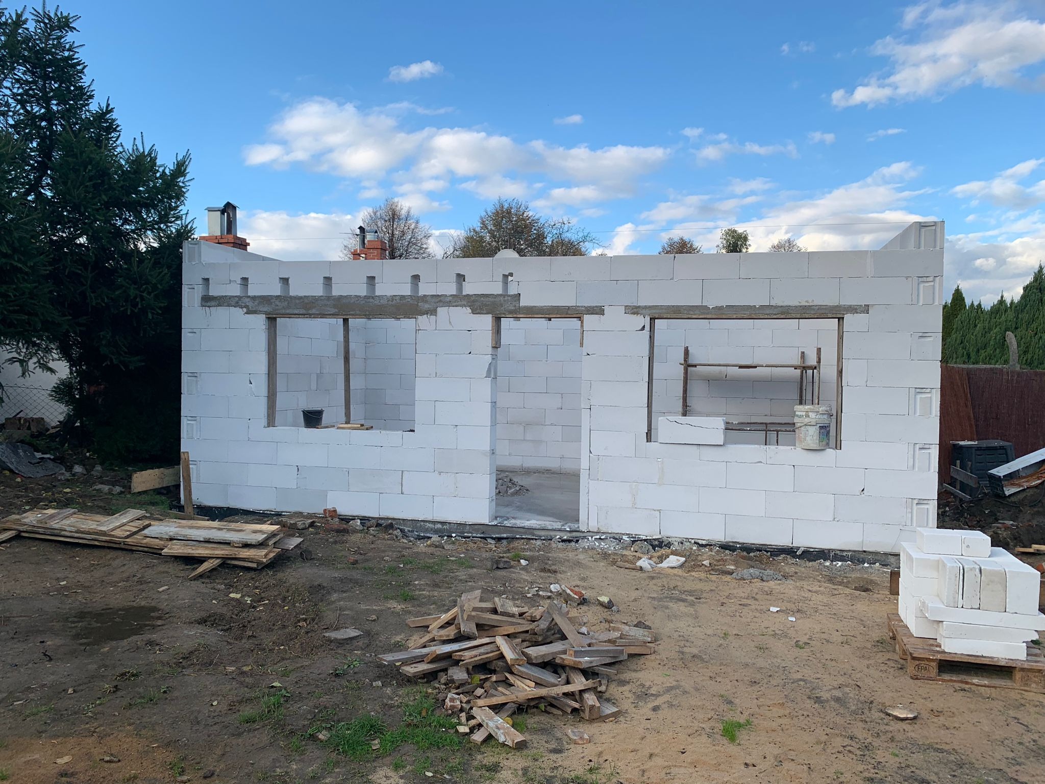 Wznoszenie ścian domu z białych bloczków, widoczne otwory okienne i drzwiowe, na ziemi porozrzucane drewniane elementy i paleta z bloczkami, w tle drzewa i niebo z chmurami.