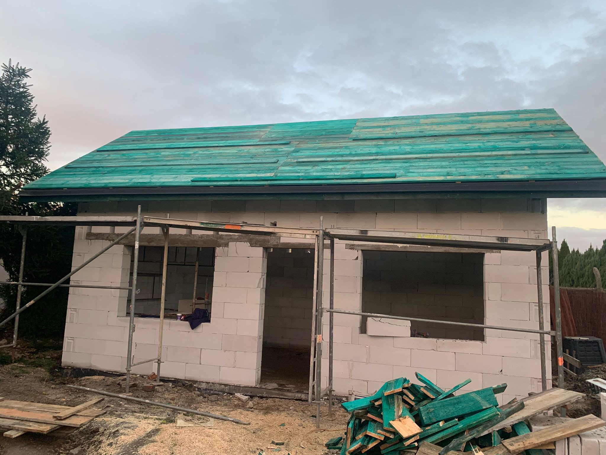 Budowa domu z pustaków: wznoszone ściany, częściowo pokryty dach deskami w kolorze turkusowym, rusztowanie, stos desek przed budynkiem.