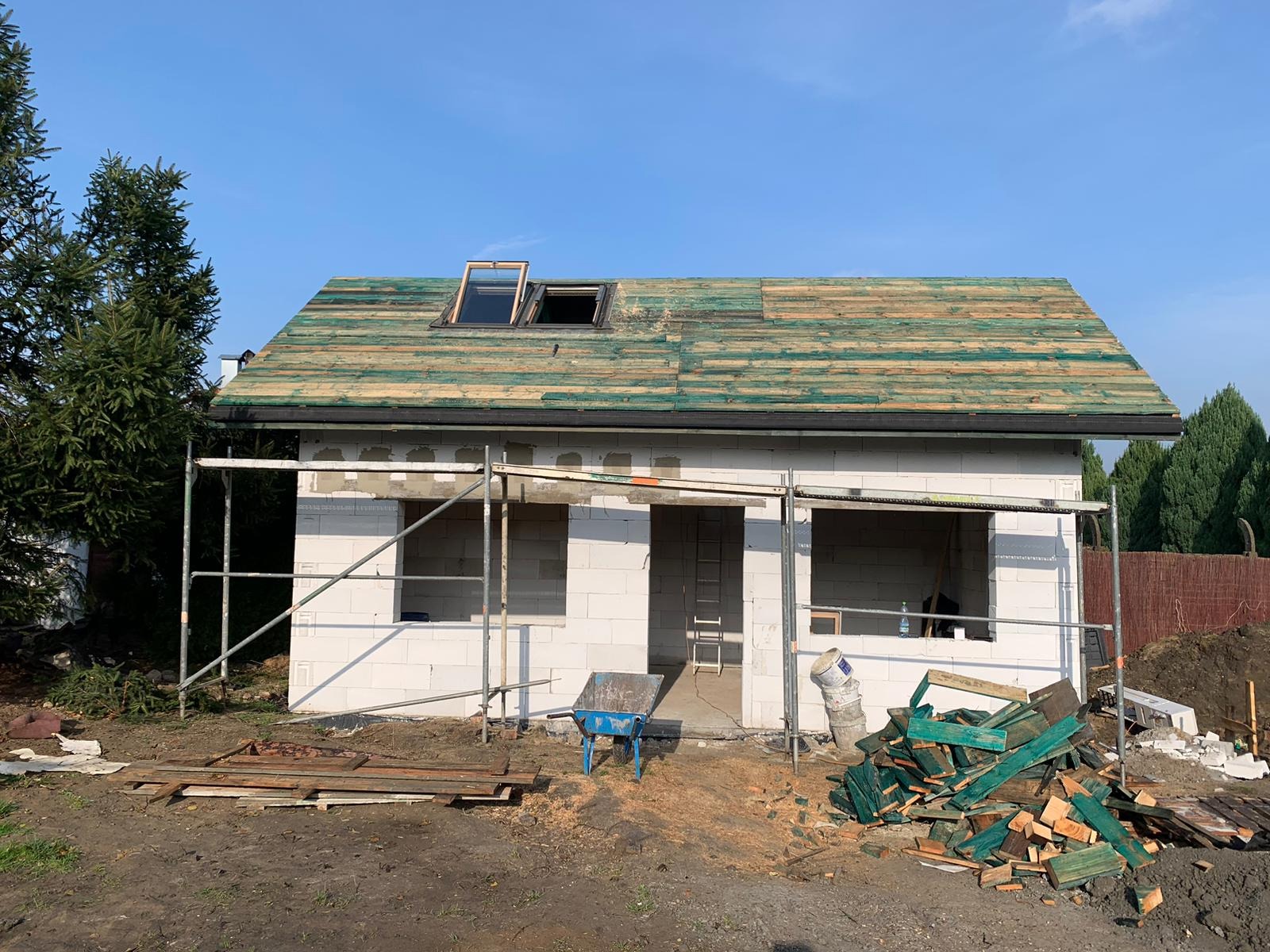 Budowa domu jednorodzinnego w trakcie realizacji: widoczne ściany z białego betonu komórkowego, drewniana konstrukcja dachu z oknami dachowymi, rusztowanie, taczka i resztki materiałów budowlanych...