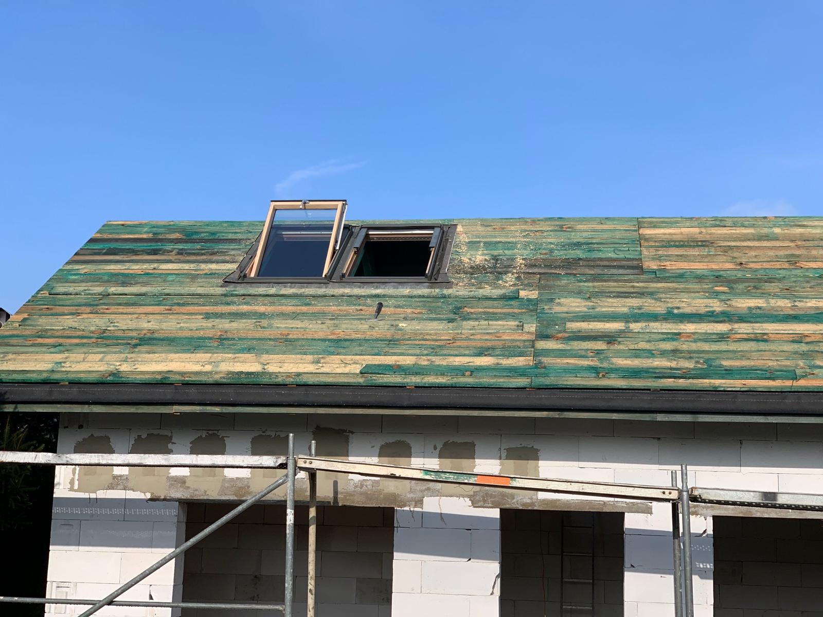 Dom w trakcie budowy z widocznym dachem pokrytym deskami z zieloną impregnacją i dwoma oknami dachowymi, jedno otwarte, drugie zamknięte. Ściany z białego bloczka, widoczne rusztowanie.