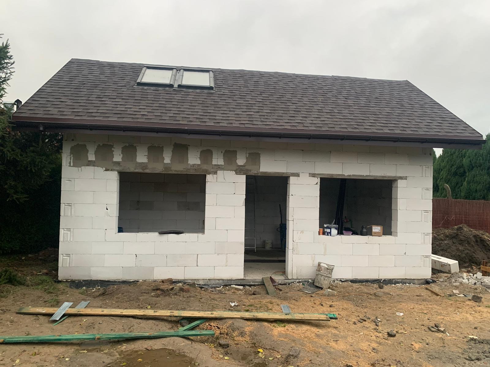 Budynek w trakcie wznoszenia z białych bloczków, widoczne otwory okienne i drzwiowe, brązowy dach z dwoma oknami dachowymi, teren wokół w trakcie prac ziemnych.
